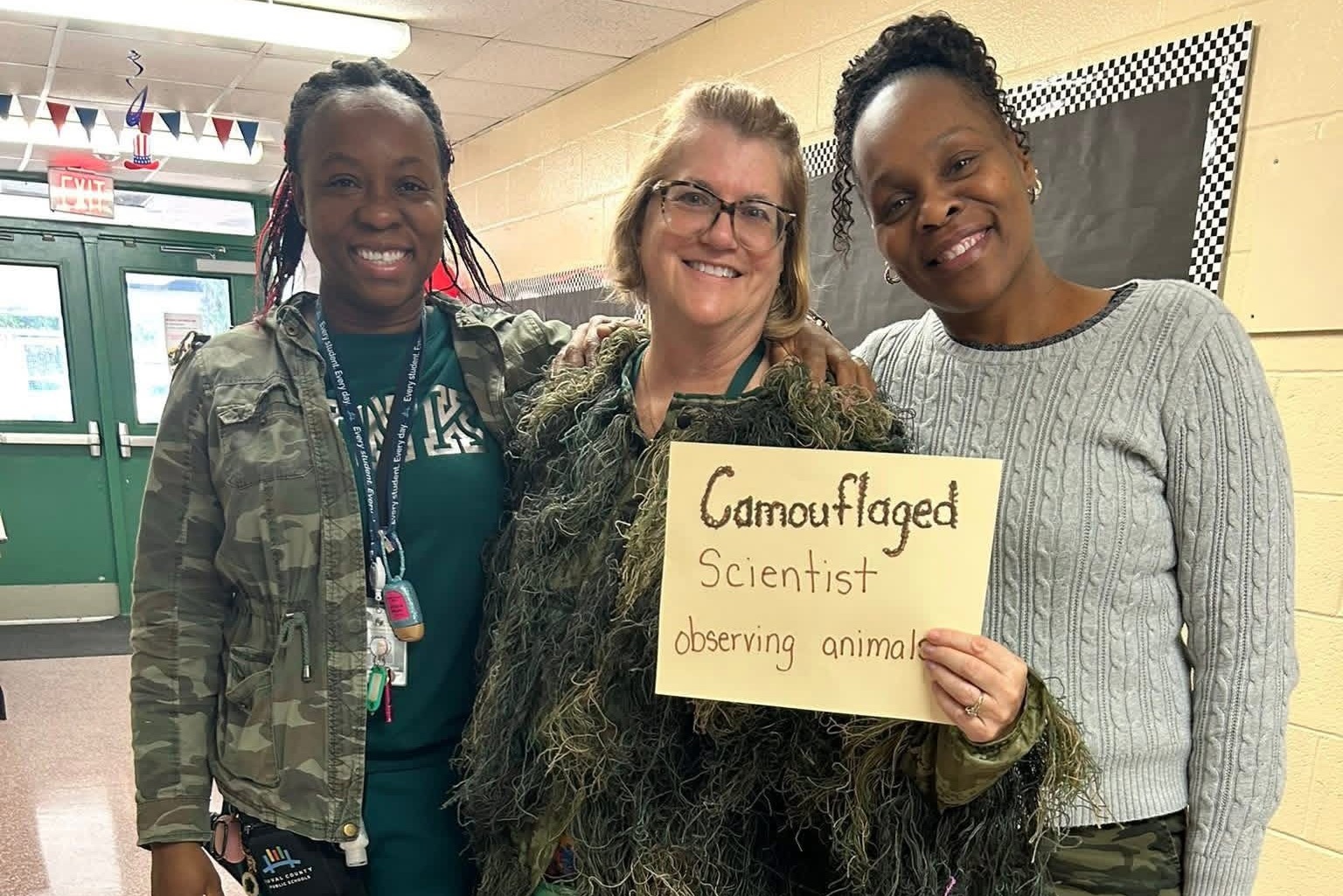 Three individuals stand in a hallway, with one holding a sign reading "Camouflaged Scientist observing animals."