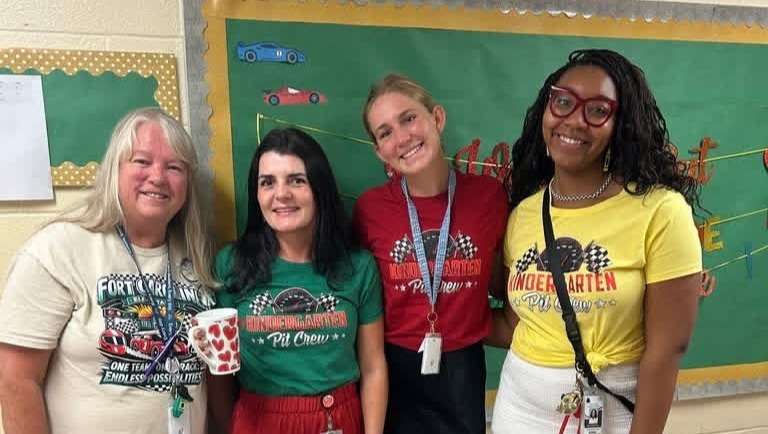 Five adults stand in front of a bulletin board. Each wears a lanyard. The woman on the left has a white shirt and black pants. The middle wears red pants. The others wear yellow and white tops.