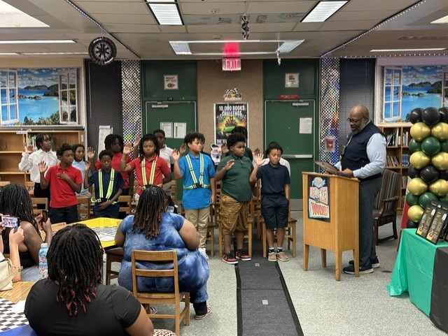 A room with a teacher and students. Students stand by a podium while the teacher speaks.