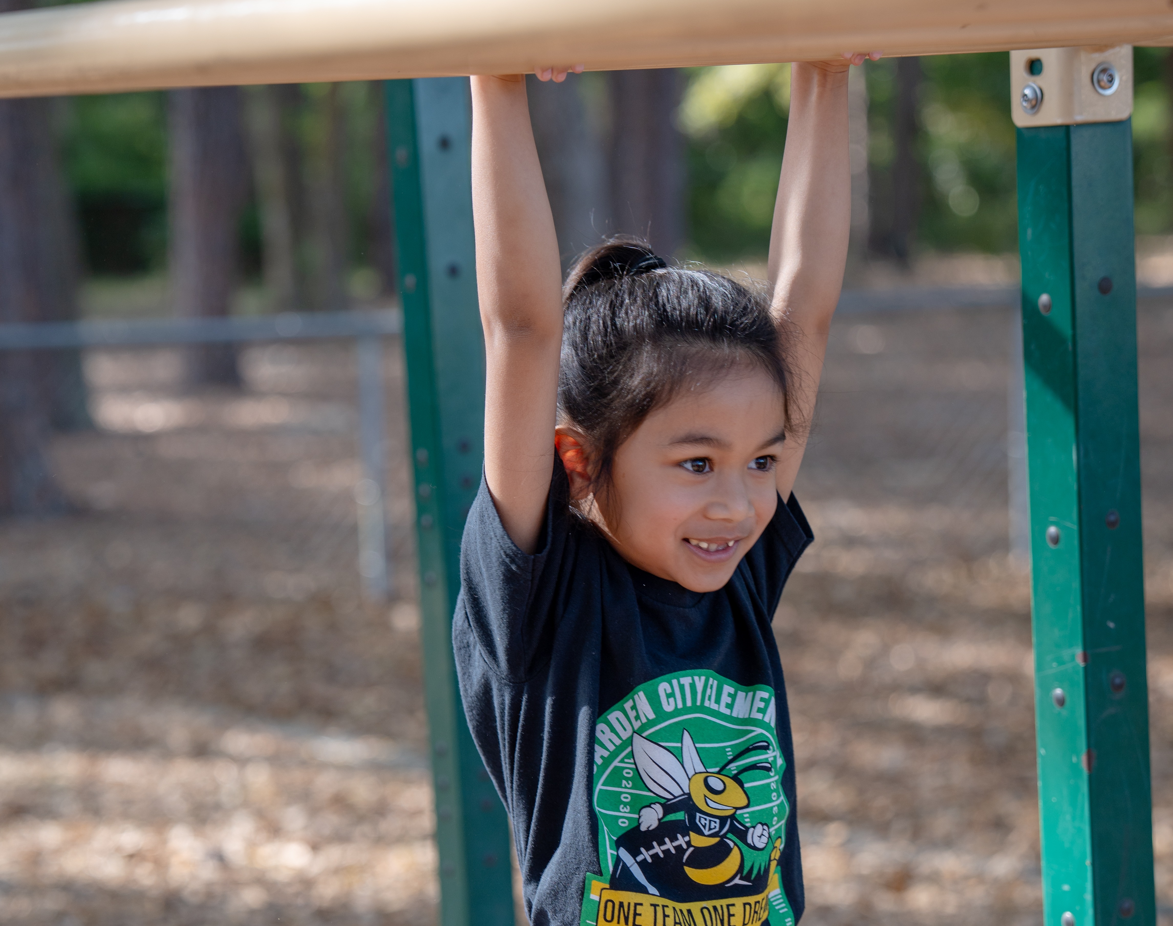 A young girl is hanging from a horizontal bar at a playground. She is wearing a black t-shirt.
