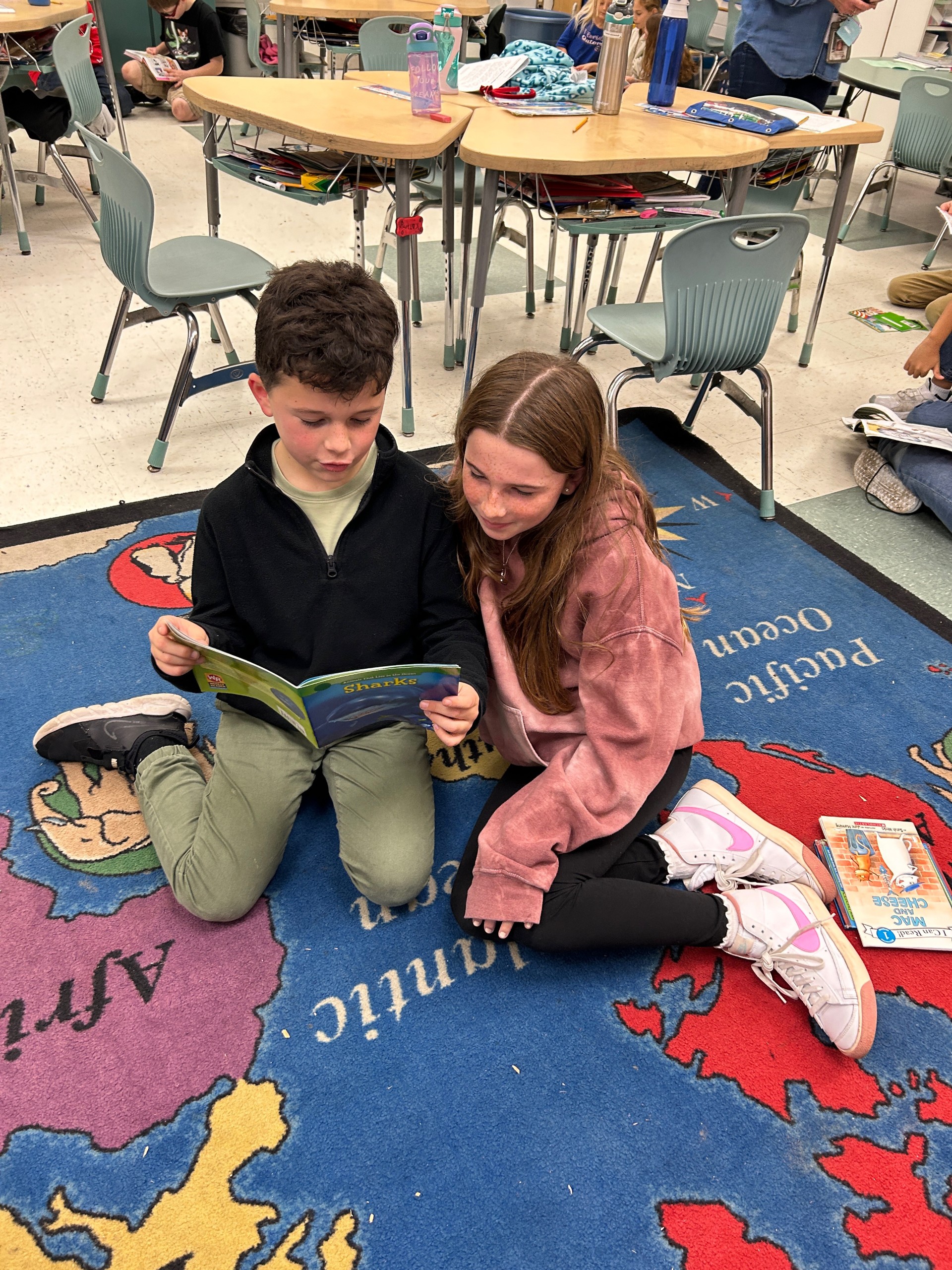 Two students, a 5th grade girl and 2nd grade boy, sitting on the floor together. The boy has a book that he is reading.