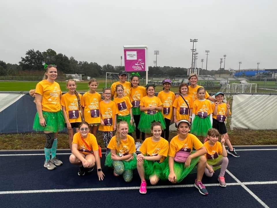 A group of people in yellow t-shirts and green skirts posing on a track field with a pink banner.