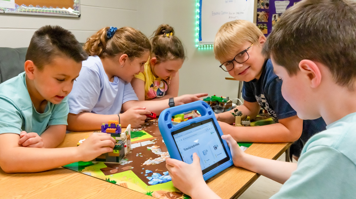 Five children sit at a table. One child holds a tablet with a colorful Lego model displayed on the screen.