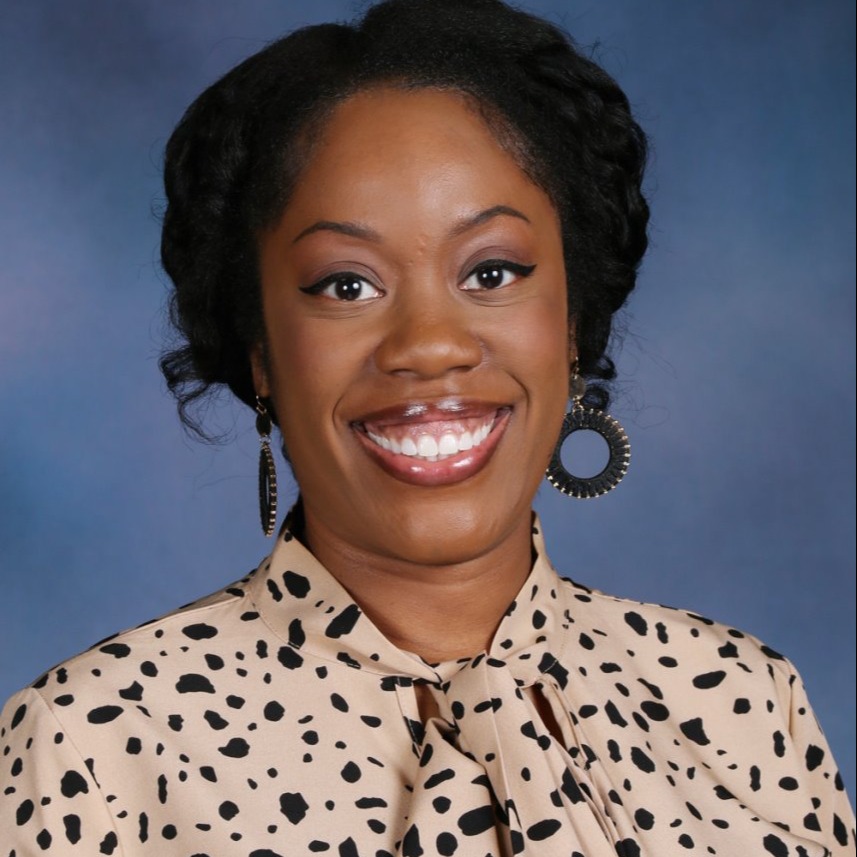 A woman with short black hair and a smile, wearing a beige blouse with black polka dots.