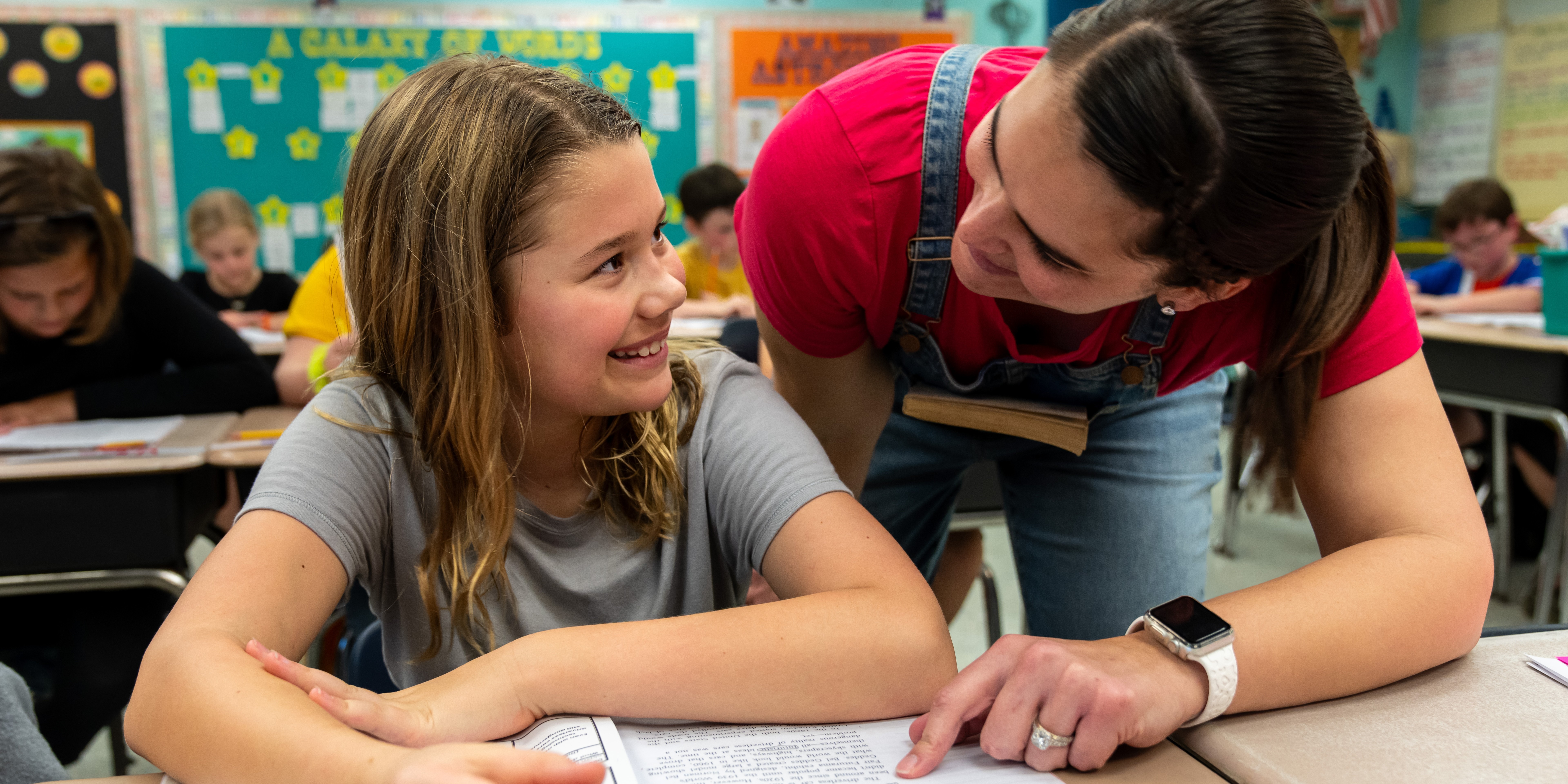 Teacher leans over desk to help student with paper; others in background seated at desks.