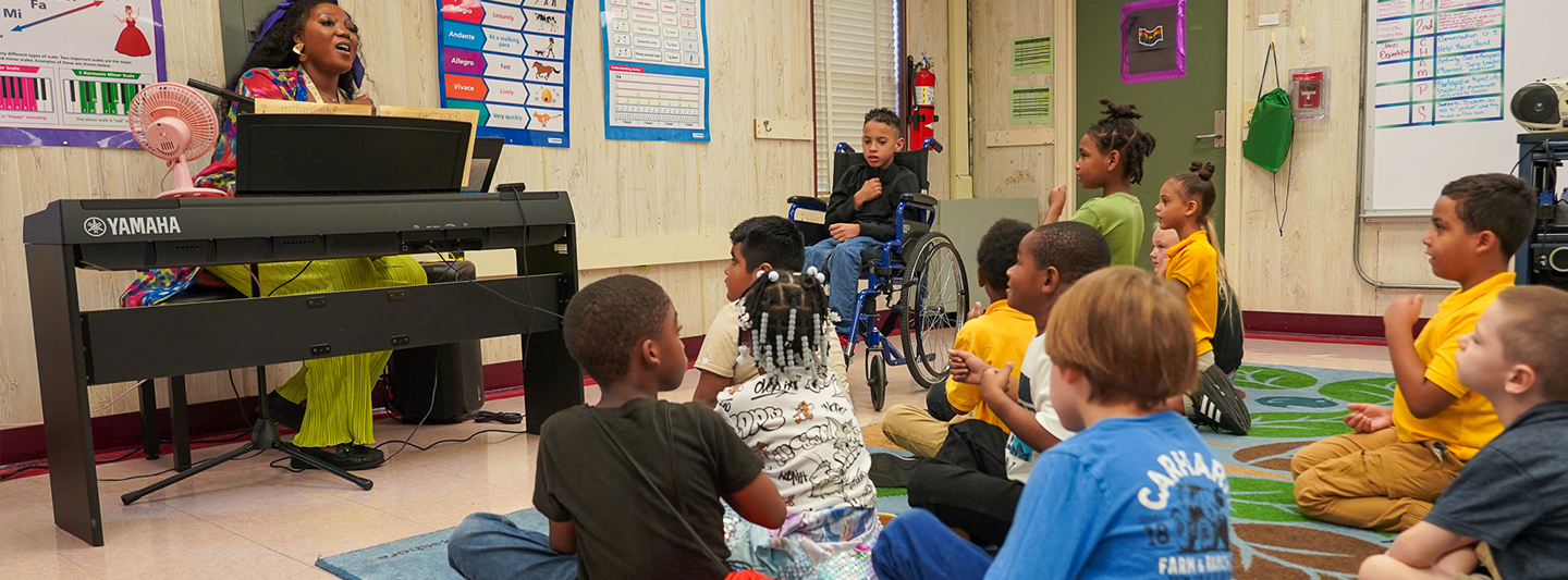 Children sitting on the floor while music teacher plays piano