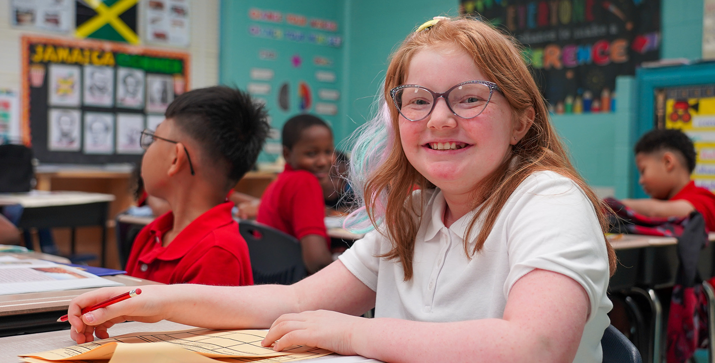 girl at her desk smiling while writing on a worksheet