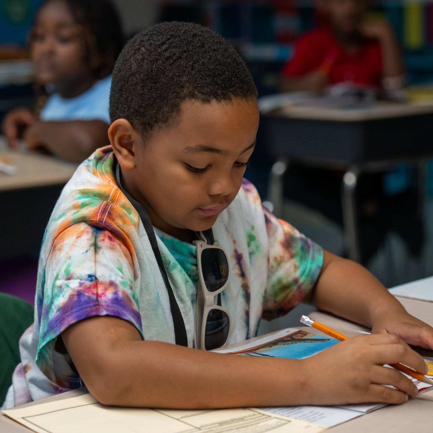 A young student sits at a desk, focused on a tablet, wearing a tie-dye shirt. Other students are also seated in the background.