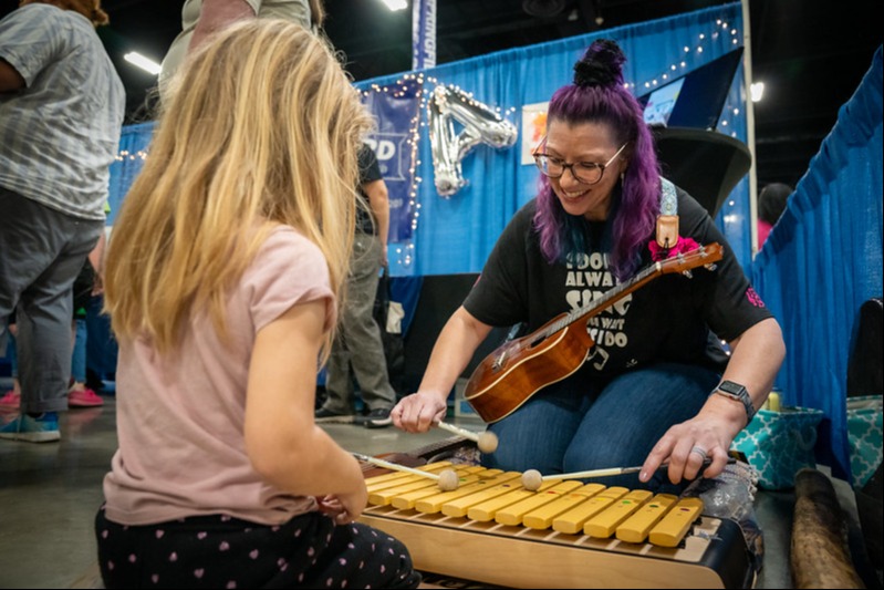 Teacher sitting on the ground holding a ukelele and playing a  orff with a student also sitting on the ground.