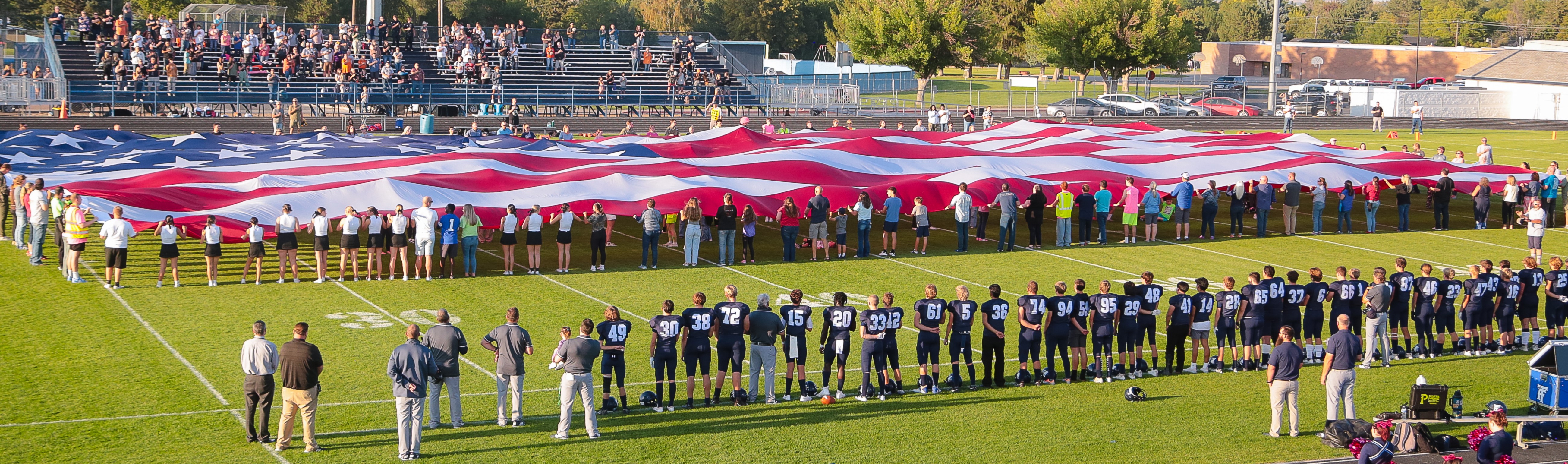 Giant American Flag on the TFHS football field with football players and coaches looking on.