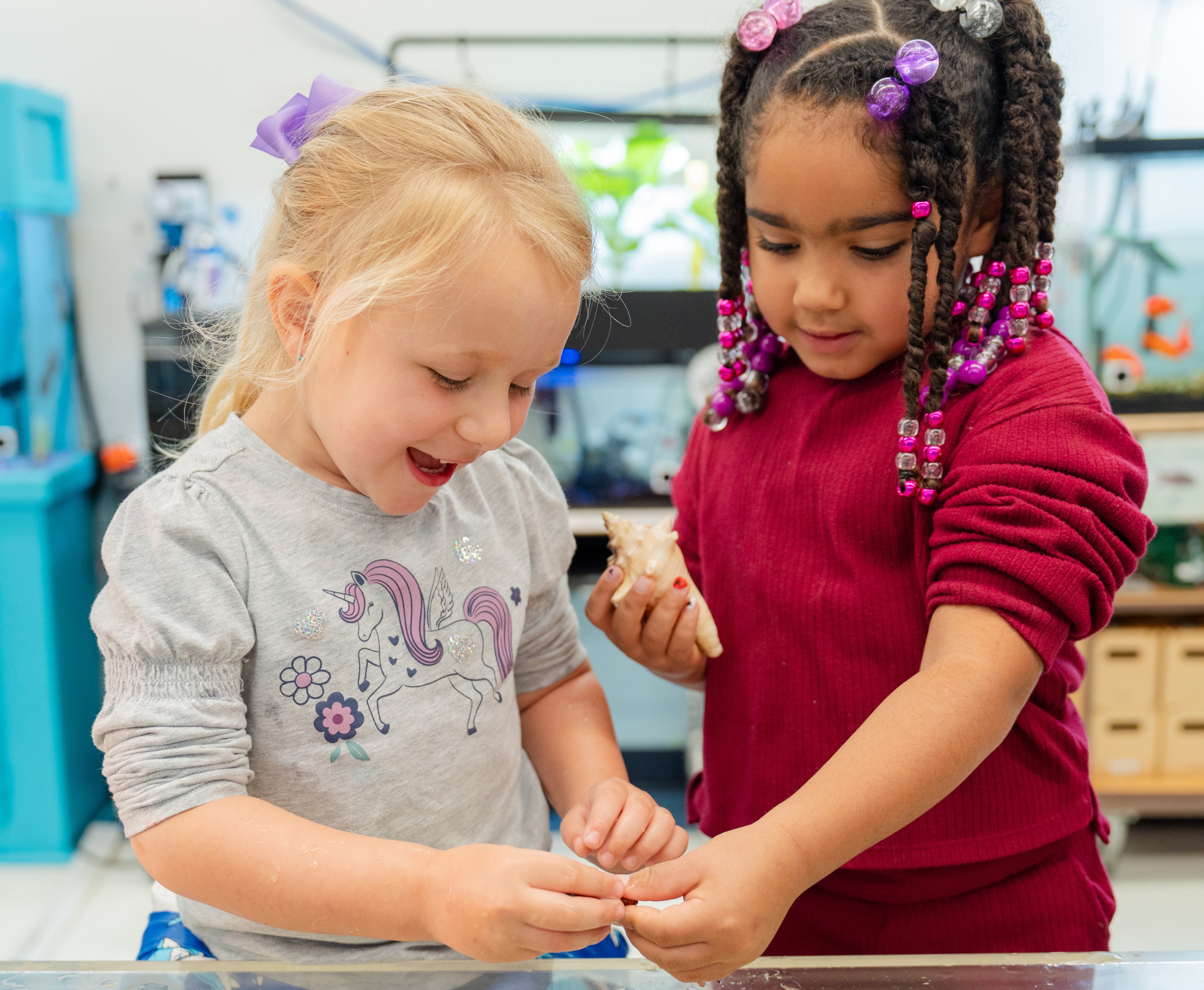 Two children examine a small object, one wearing a gray shirt with a unicorn, the other in a red top.
