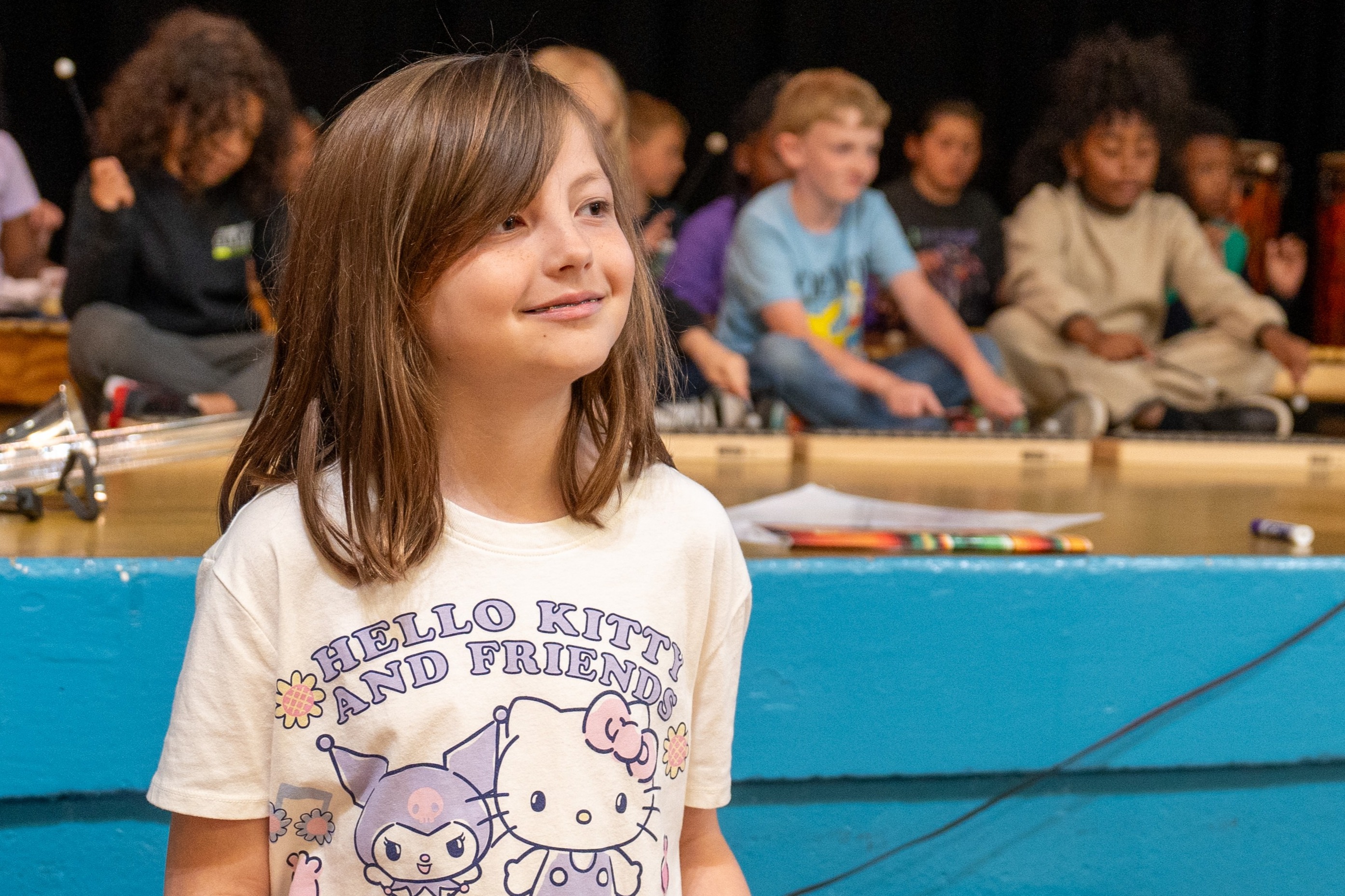 A girl plays a xylophone in front of a blue wall, with other children seated on the floor.