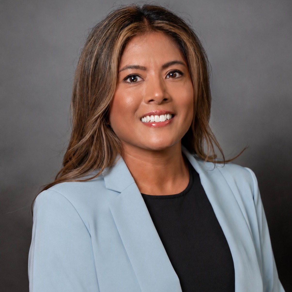 A smiling woman in a light blue blazer and black top stands against a gray background.