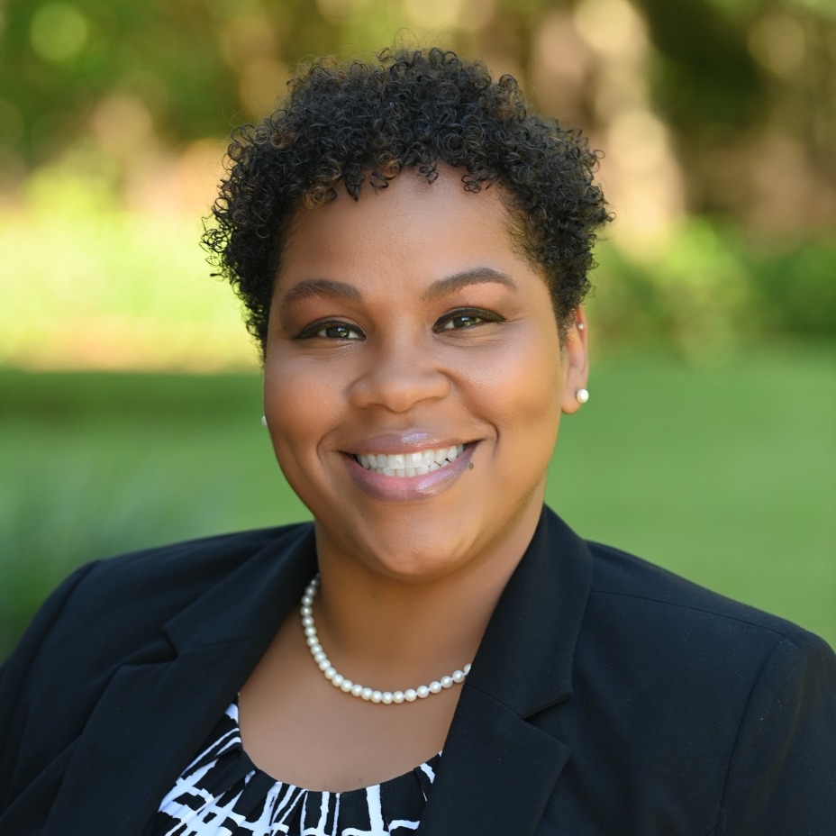 A woman with short curly hair stands outside, smiling. She wears a black blazer over a white and black patterned blouse and a pearl necklace.