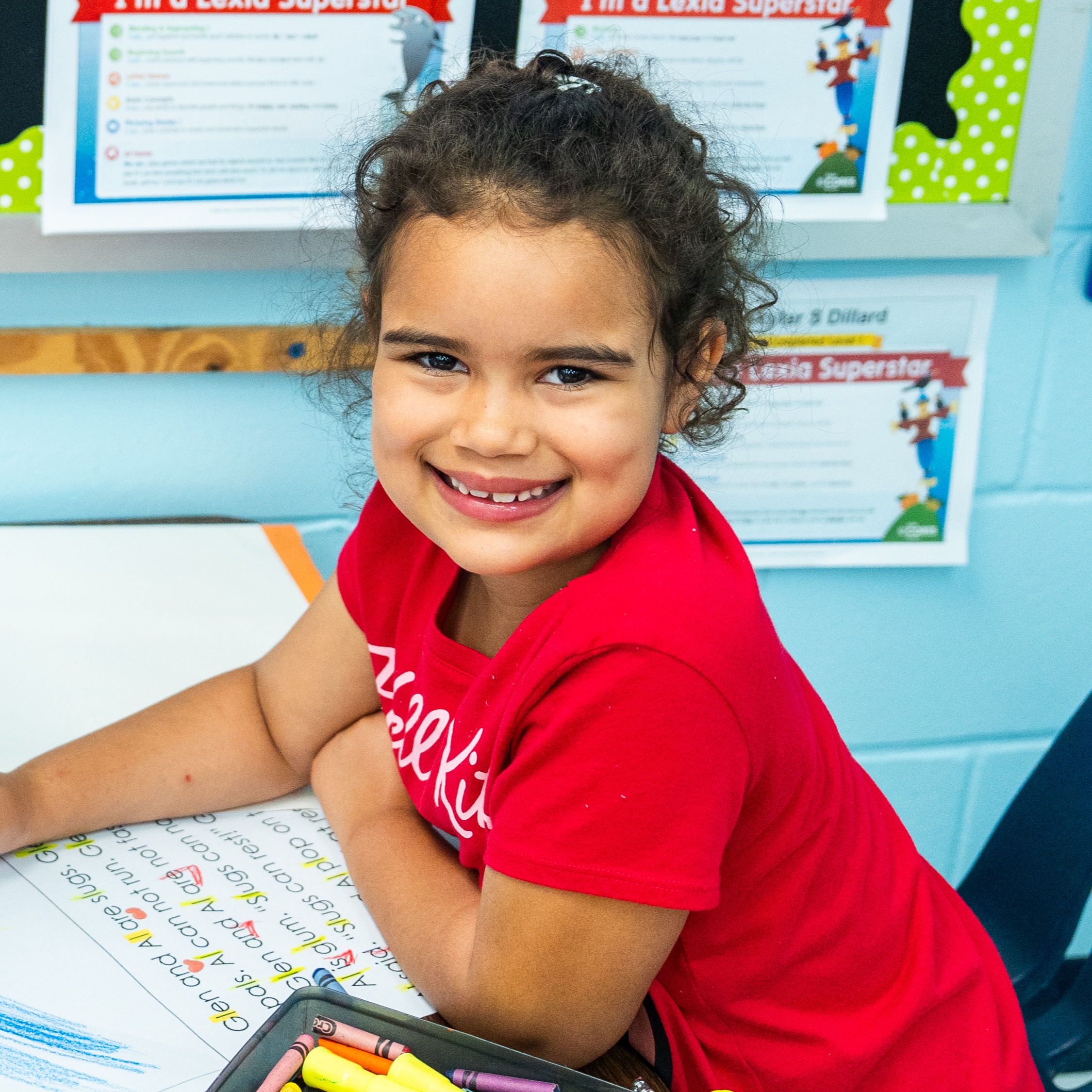 A smiling child in a red shirt sits at a desk, drawing with a crayon on a large piece of paper.