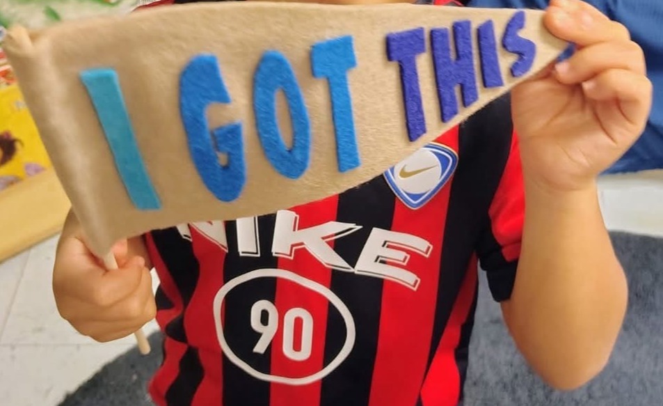 A young child in a red and black striped shirt holds a sign that says "I GOT THIS".