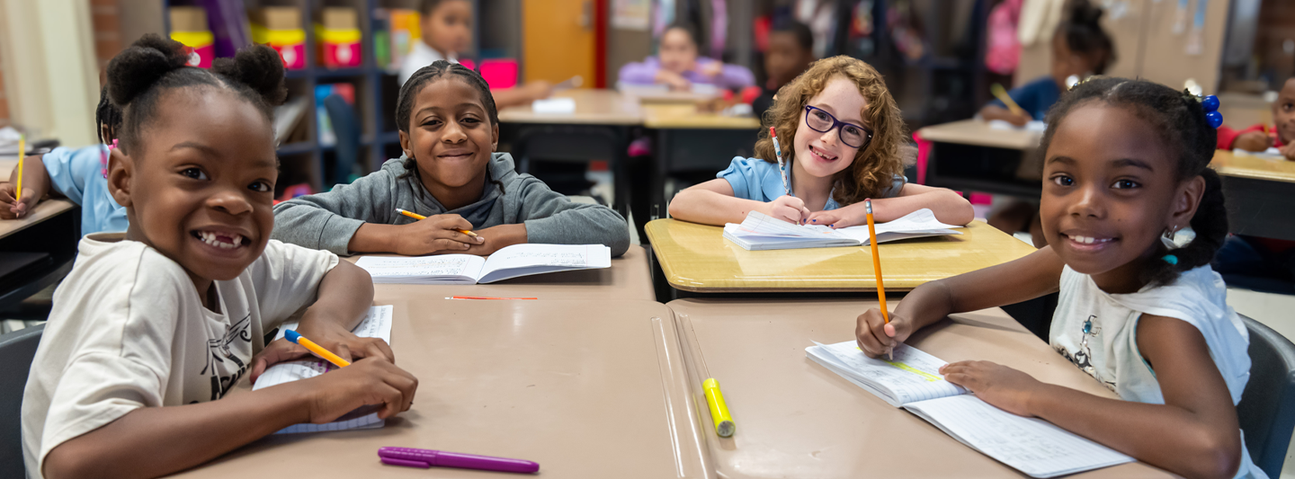 smiling students in a desk pod writing in their notebooks