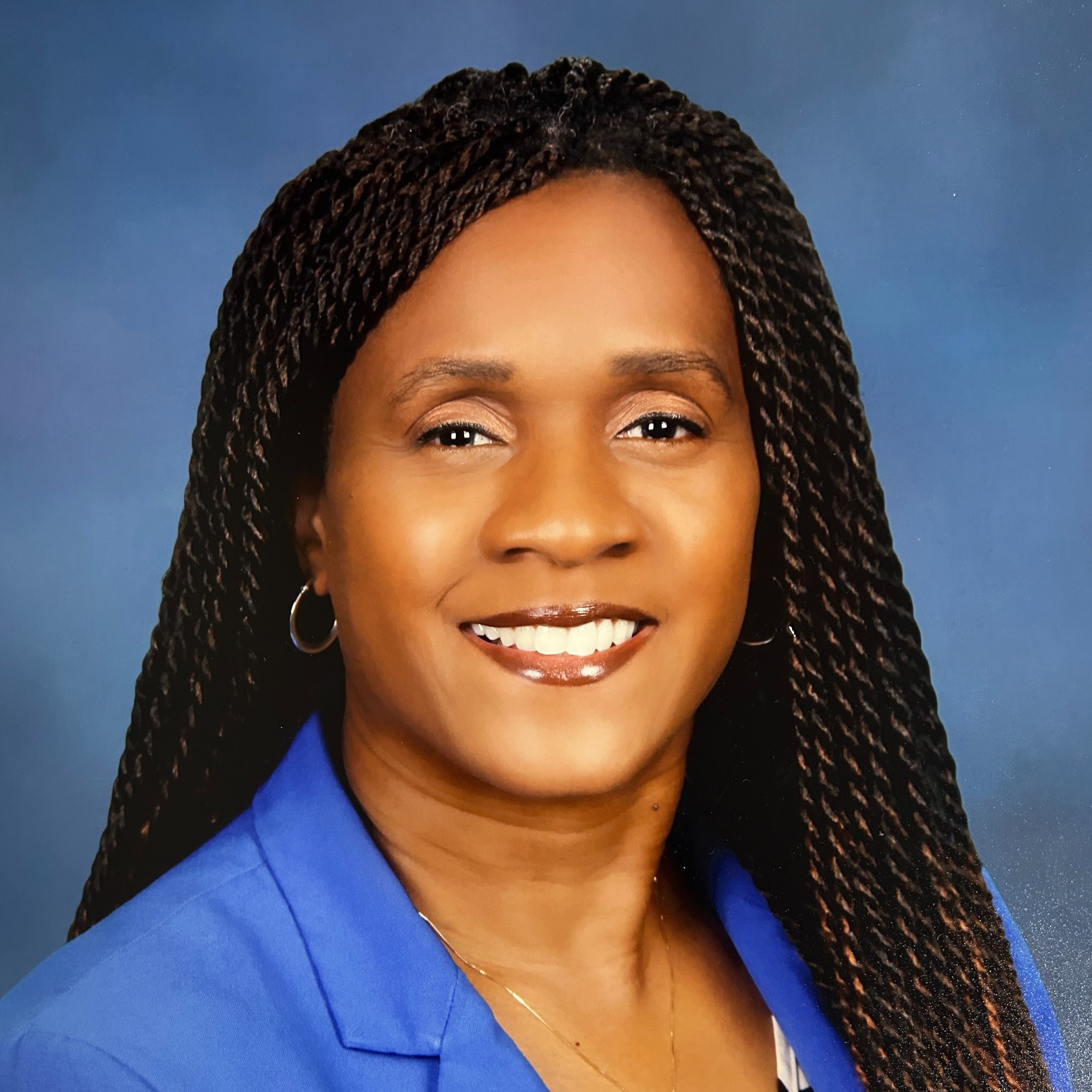 A woman with long, braided hair wearing a blue blazer, striped shirt, and necklace, smiling, and standing against a blue background.