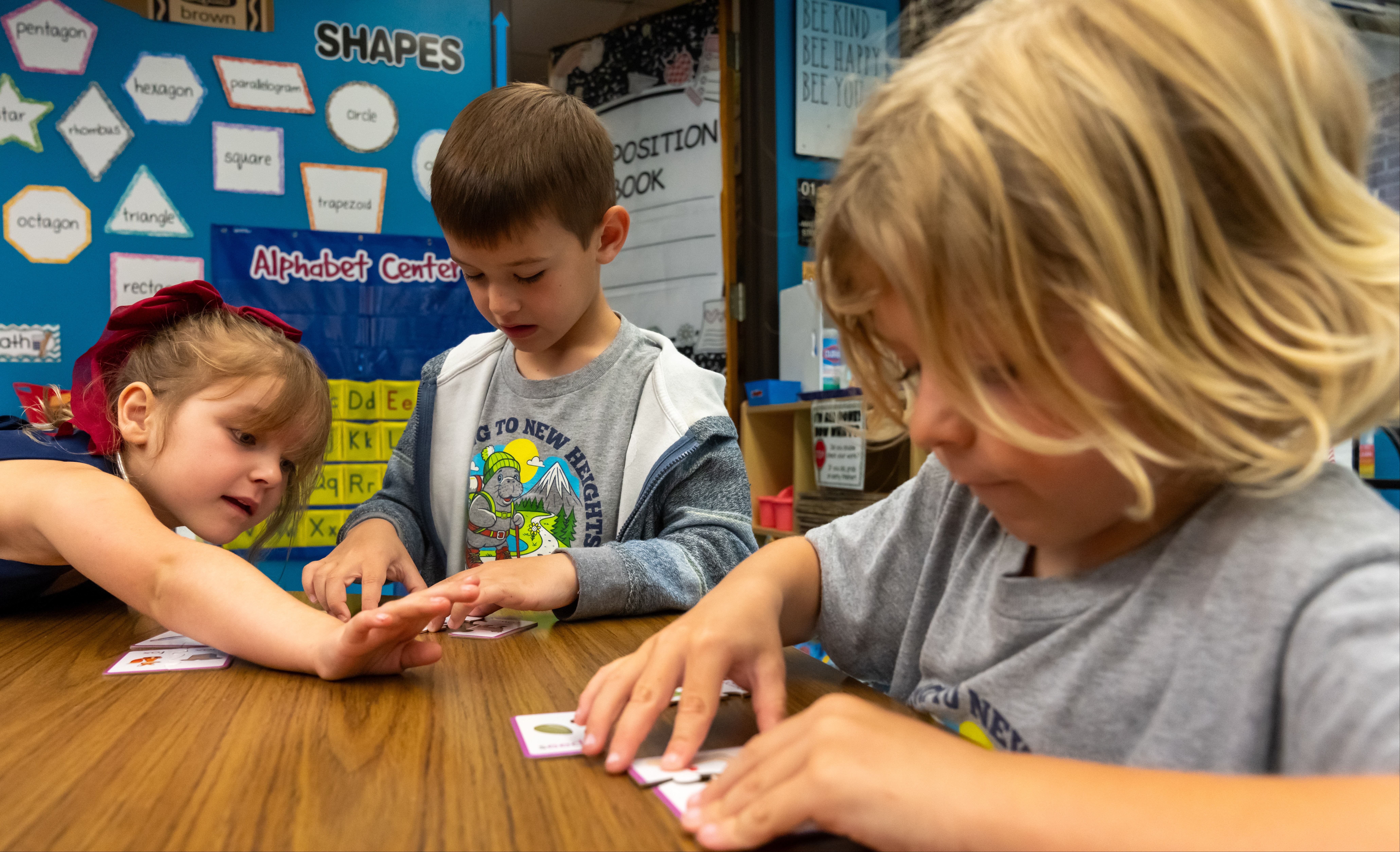 Three children collaborate at a table, with one reaching out to another. Background contains educational posters.