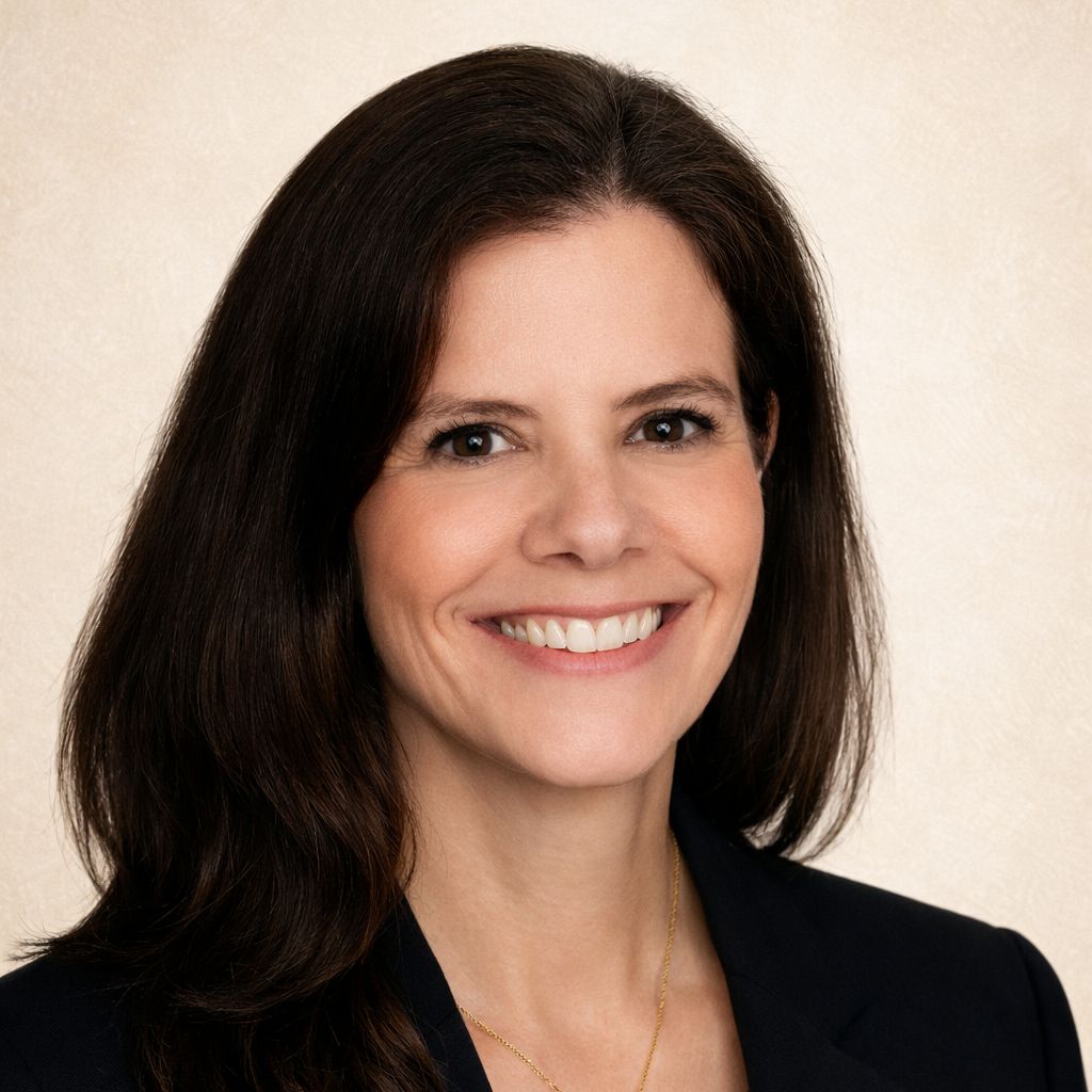 Person wearing a black blazer, white top, and gold necklace, smiling and looking forward with a beige background.