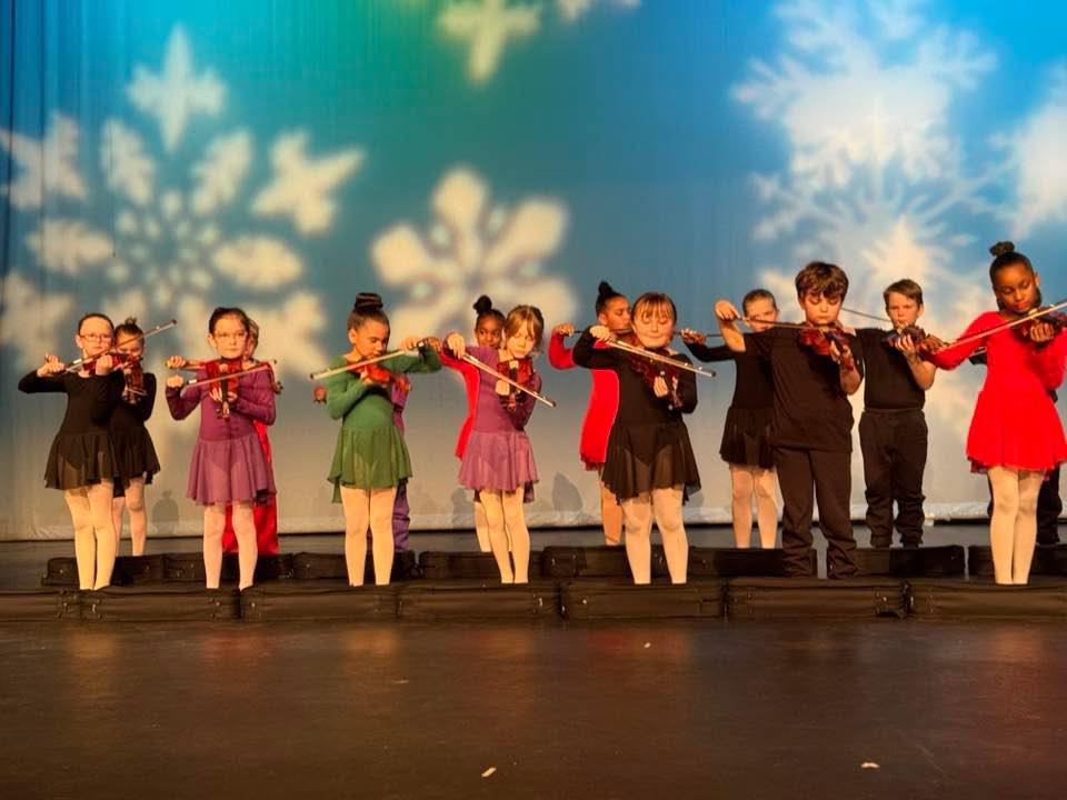 A group of children on stage playing violins; snowflake designs on a blue and green background.