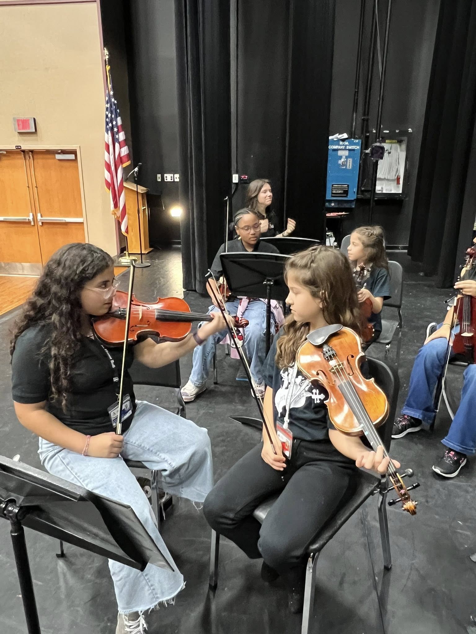 Two girls seated in chairs, holding violins. One plays while the other listens. Music stands, a flag, and other chairs in background.