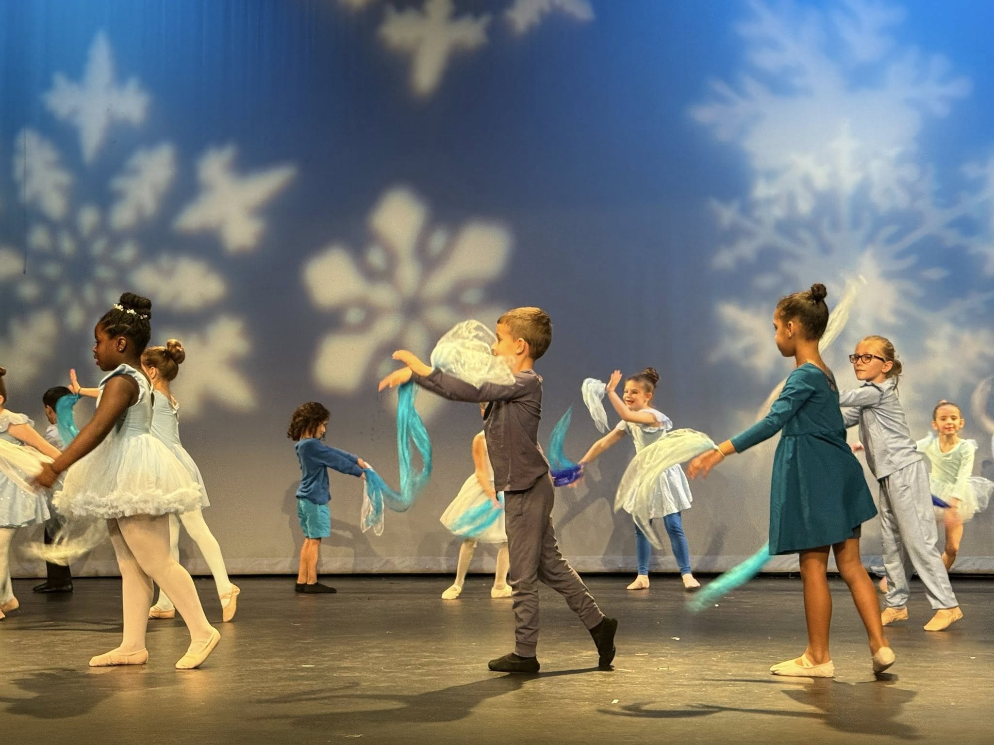 Several children perform a ballet on a stage with snowflakes in the background. Some hold ribbons.