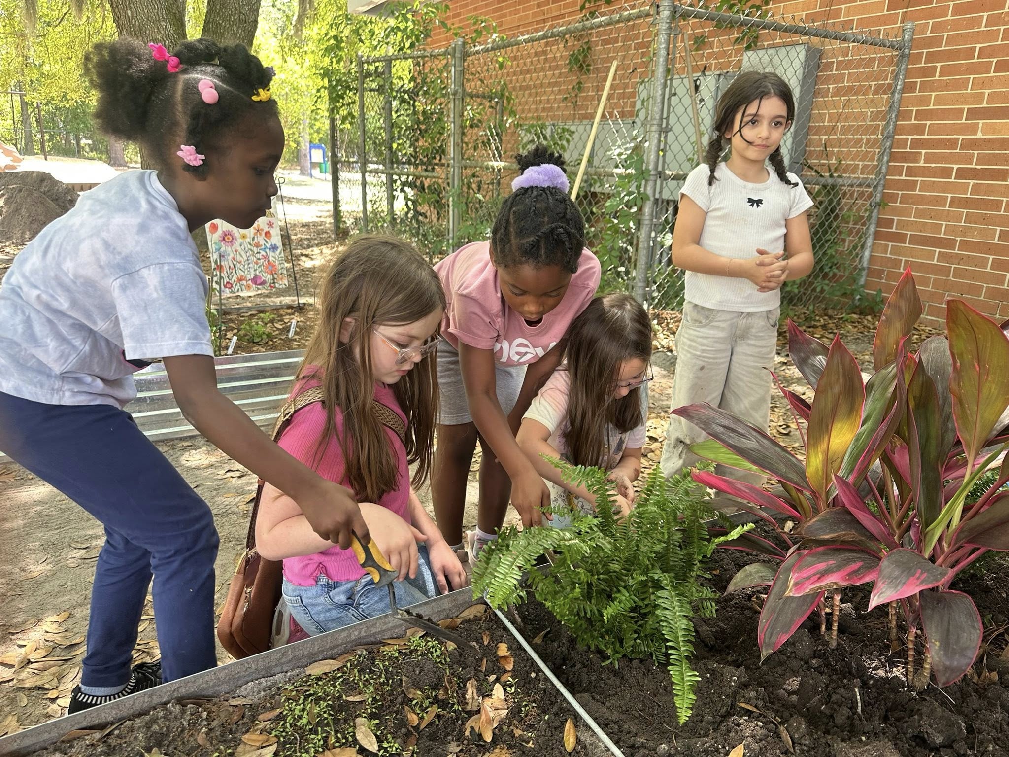 Five children in a garden, using tools to plant. Brick wall behind them.