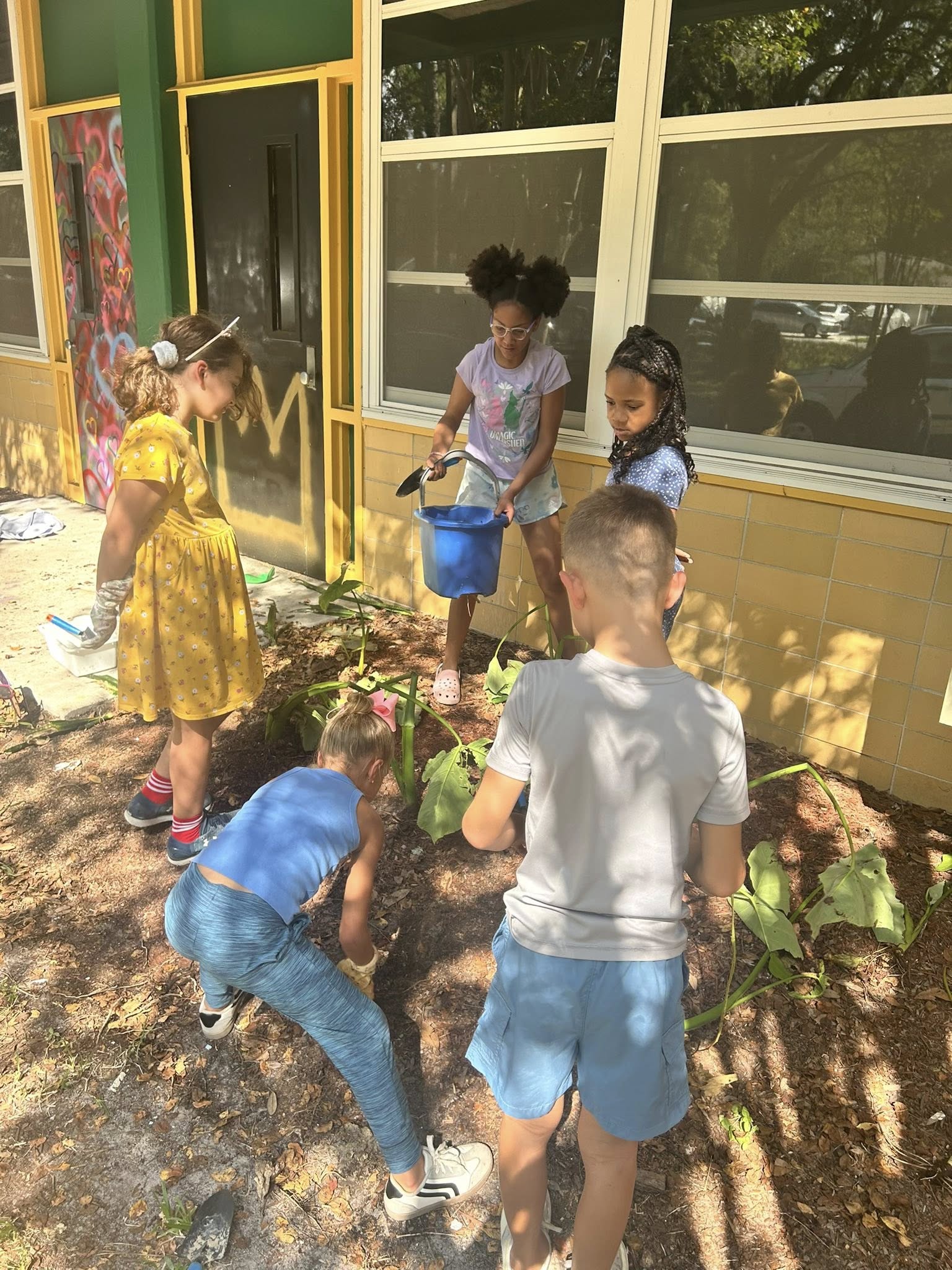 Five children plant and water plants outside a building. One child holds a bucket.
