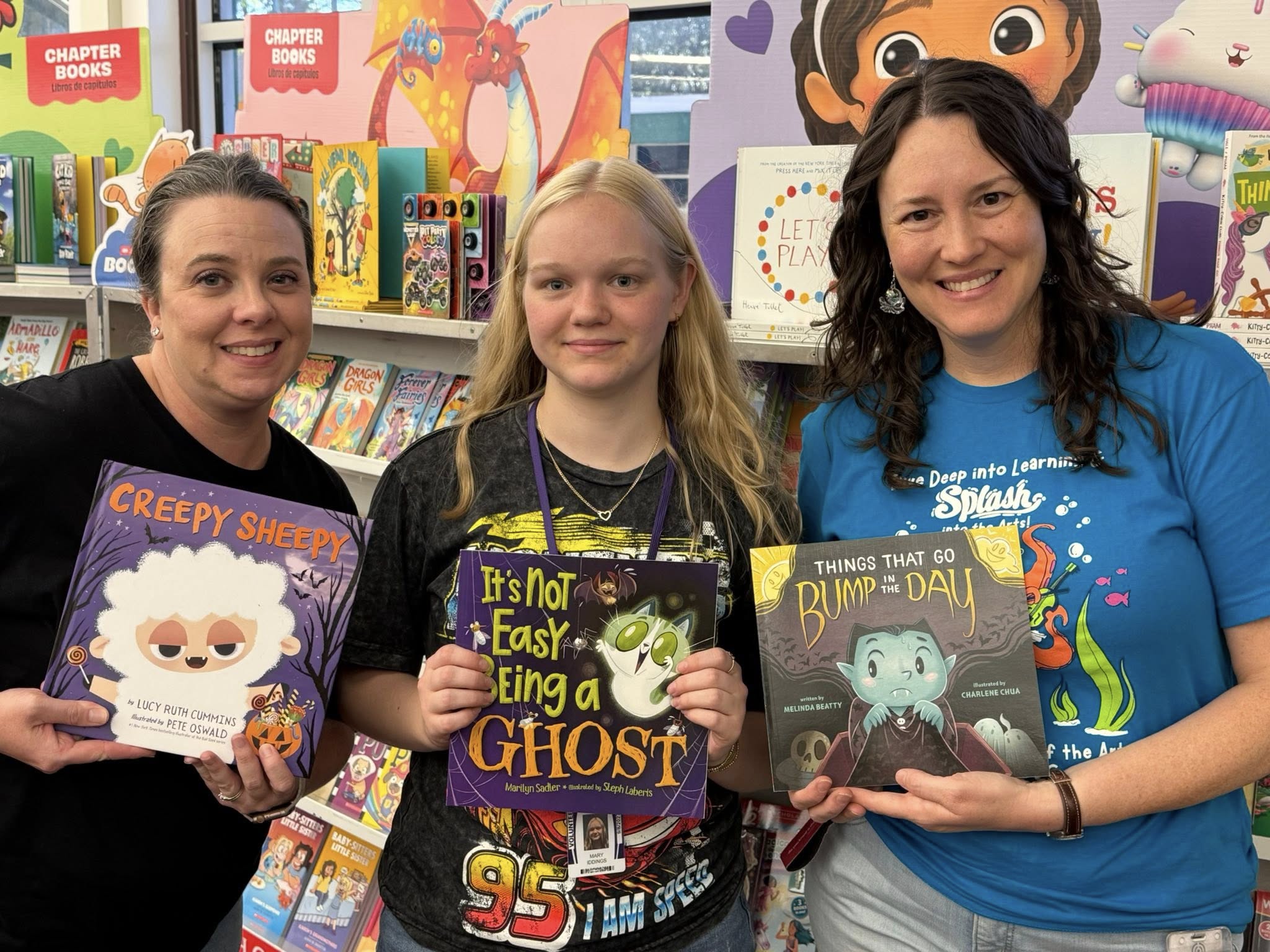 Three women stand together in a bookstore holding books. One holds a book titled "Creepy Mysteries."