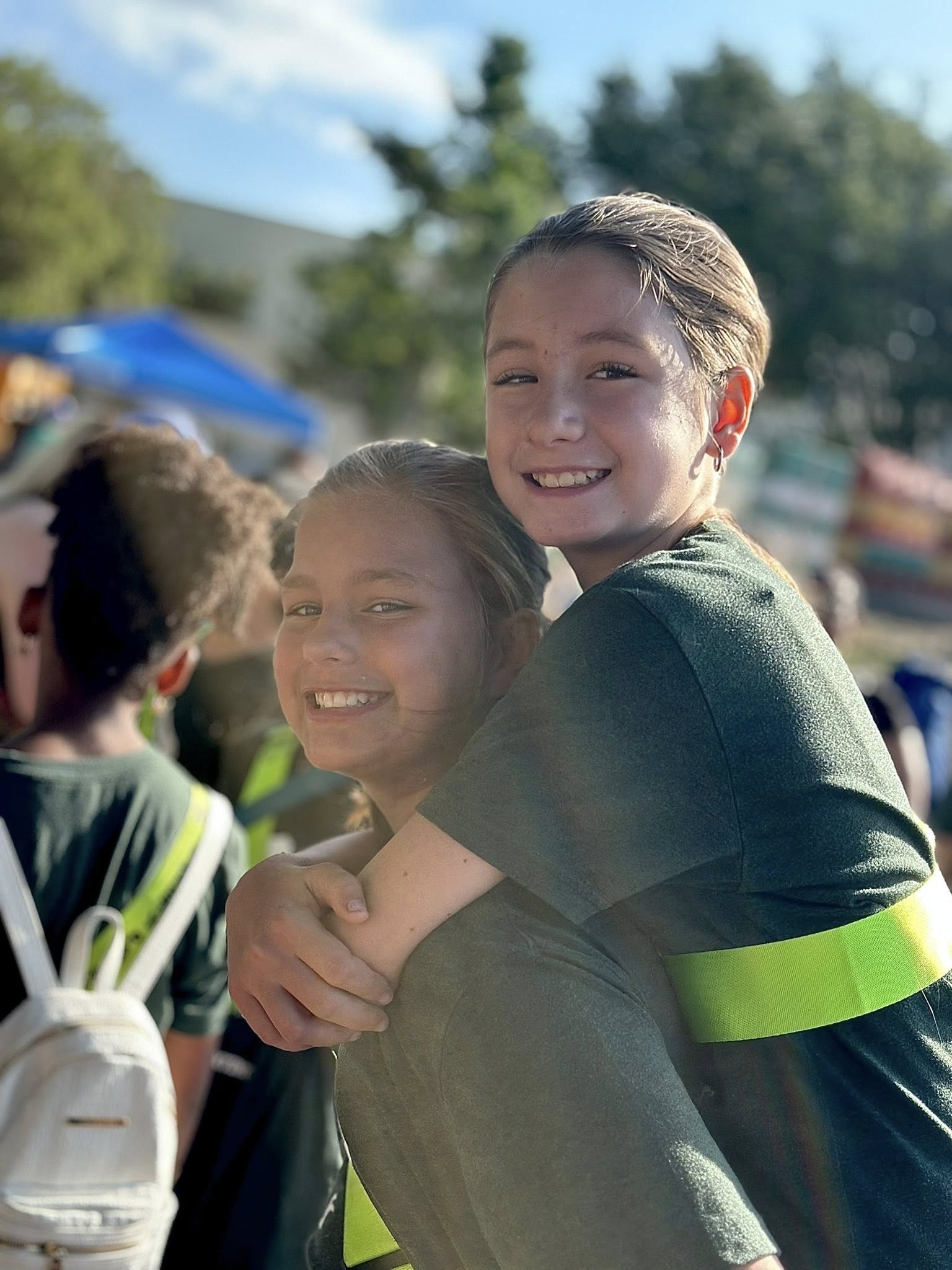 Two children in green shirts, one hugging the other, with a blurred background of a school event.