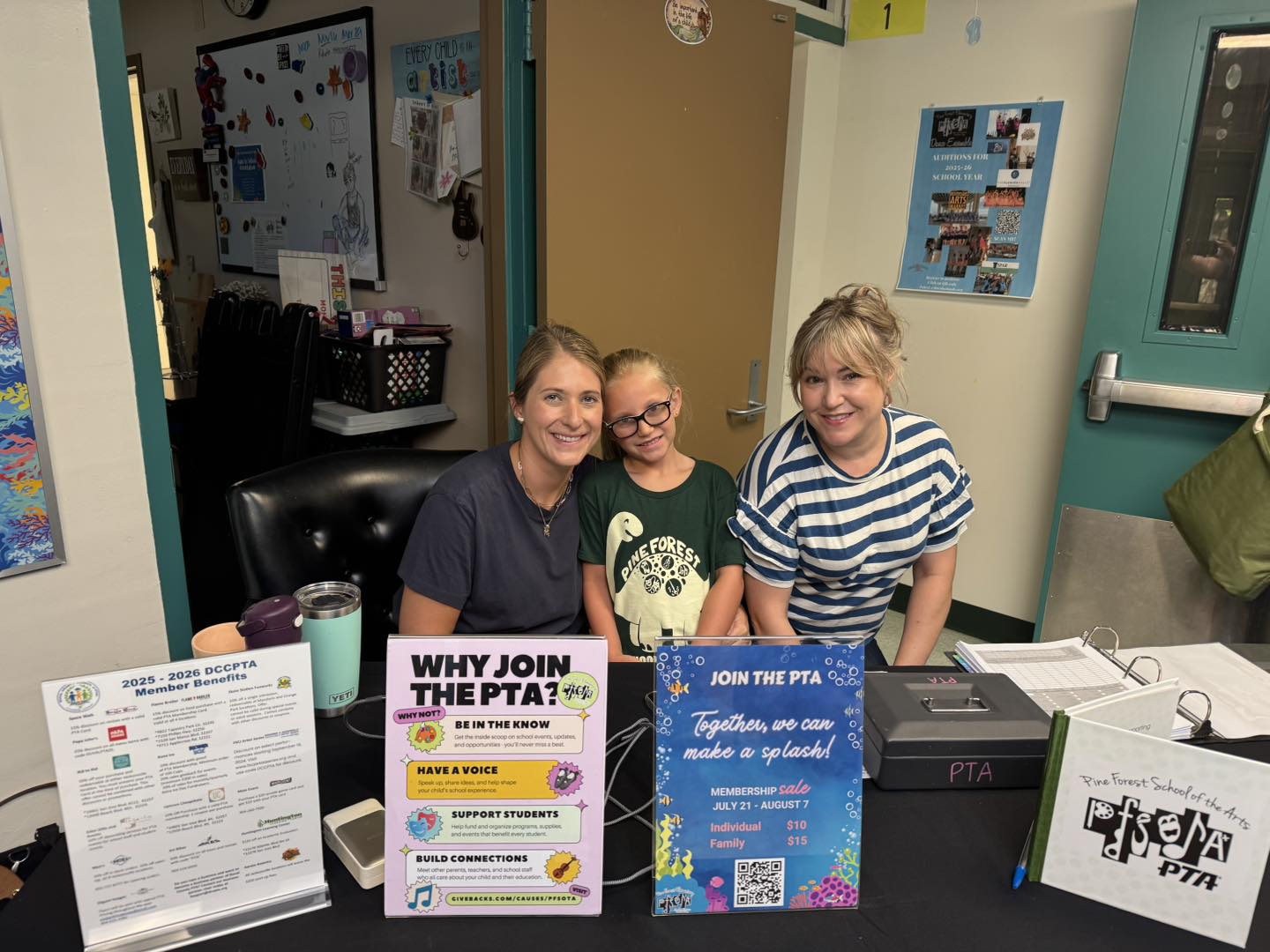 Three individuals at a table with promotional materials, including signs. A black desk holds cups, pamphlets, and a computer.