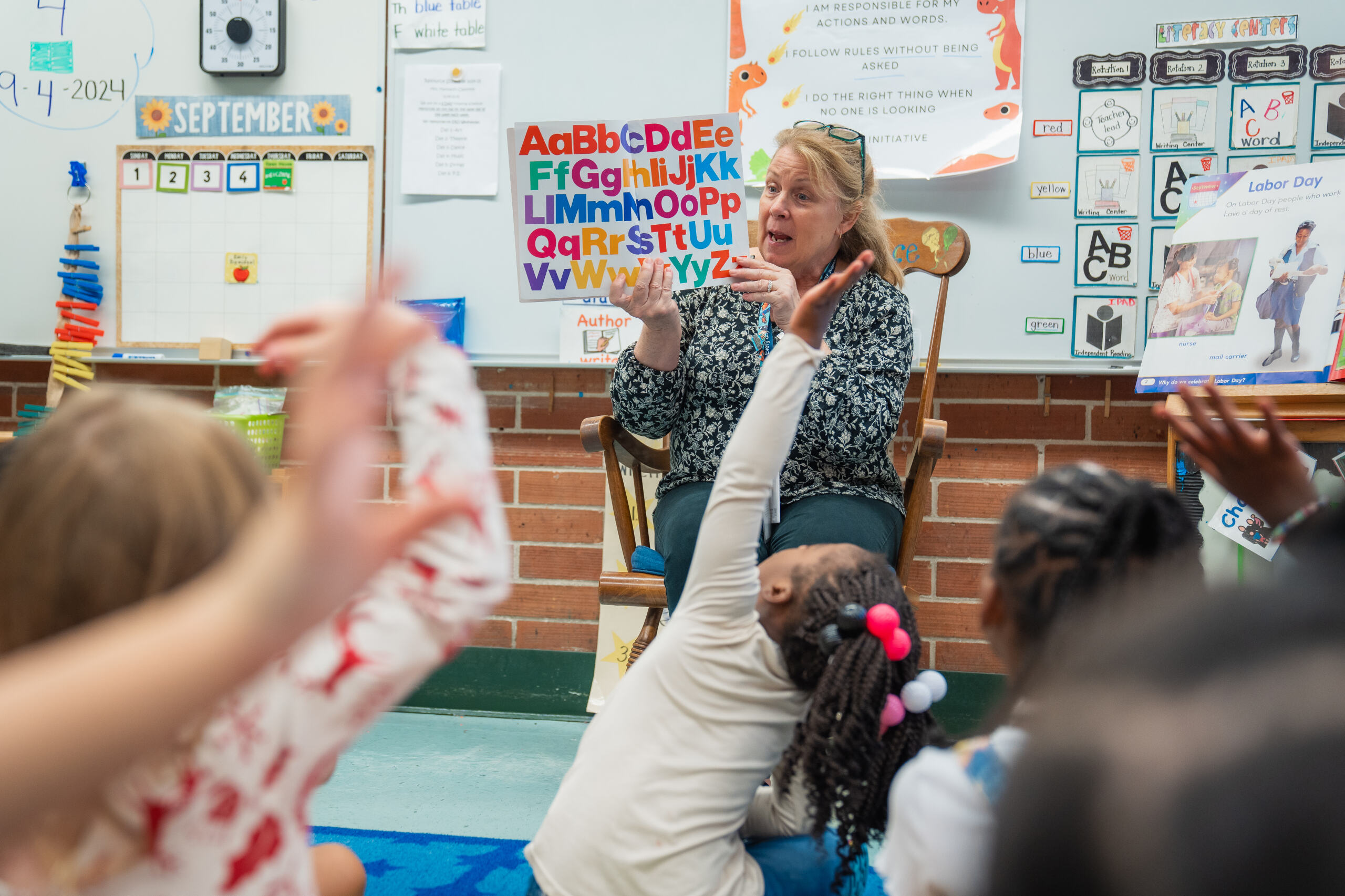 A teacher reading a book to students raising their hands on a carpet