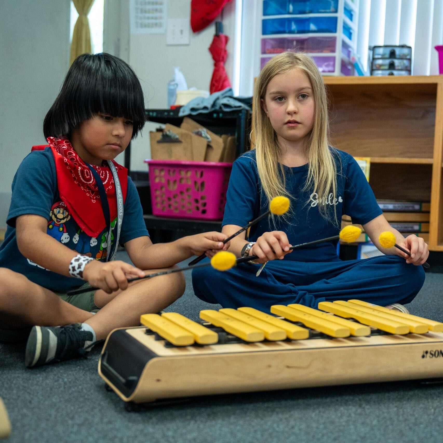 Two students sitting on the carpet playing an instrument together