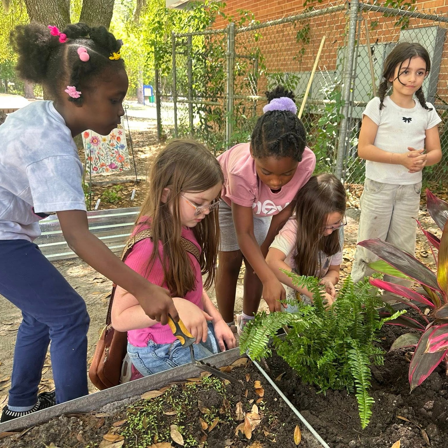 Five girls in a garden with a brick wall in the background; they are interacting with plants.