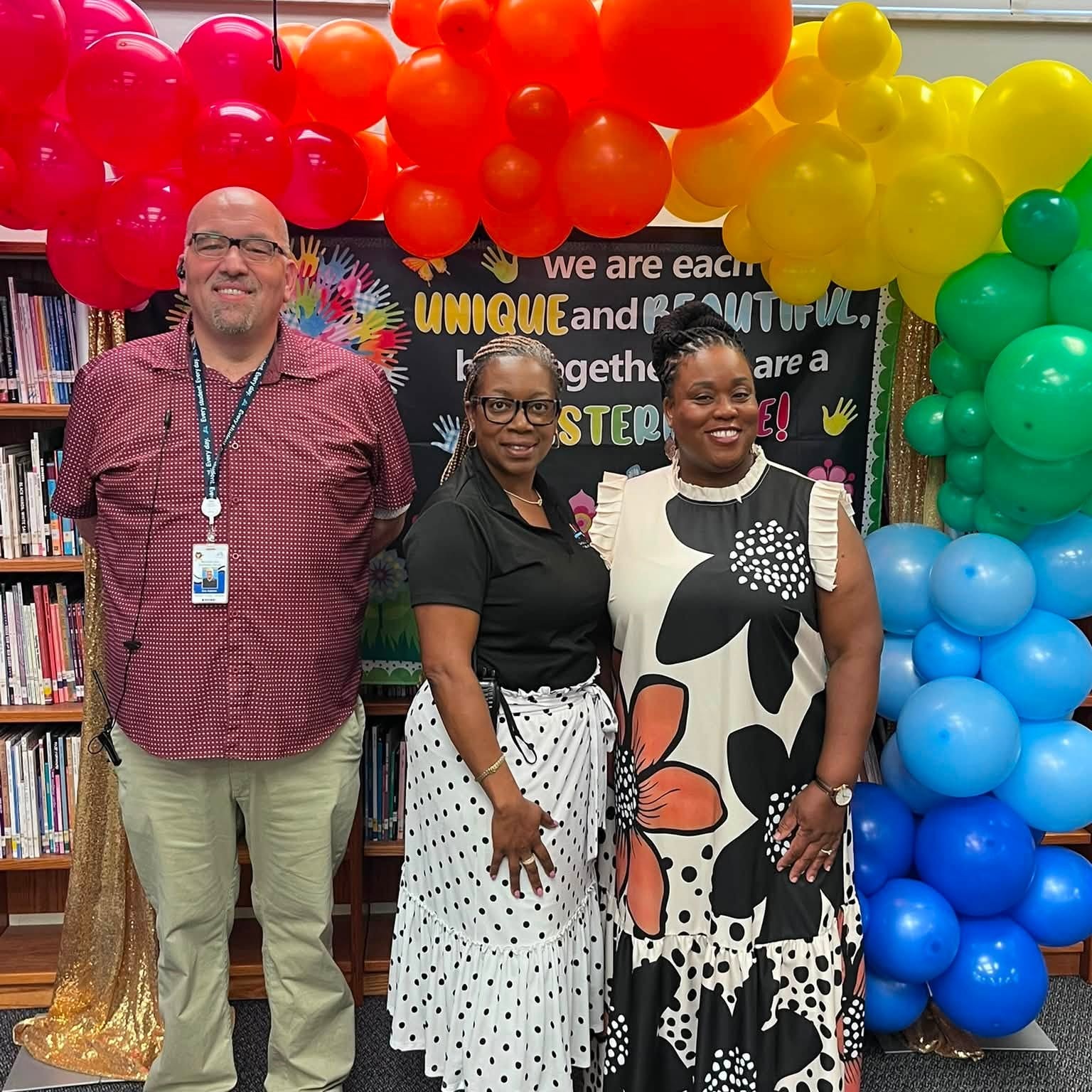 Three individuals stand smiling in front of a library backdrop with colorful balloons and a banner reading, "we are each unique and powerful together."