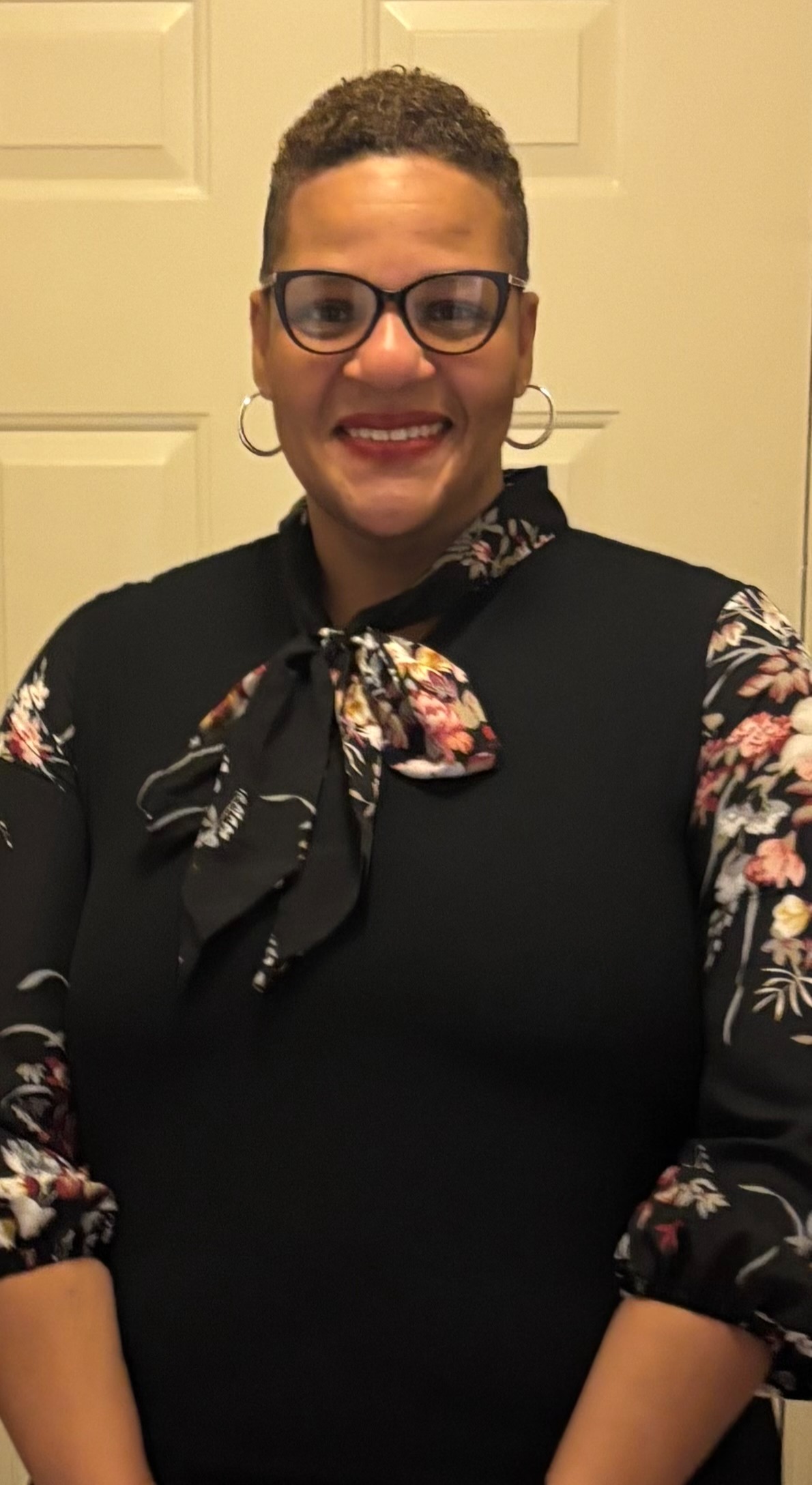 Woman with glasses, short hair, and a black floral blouse standing in front of a white door.