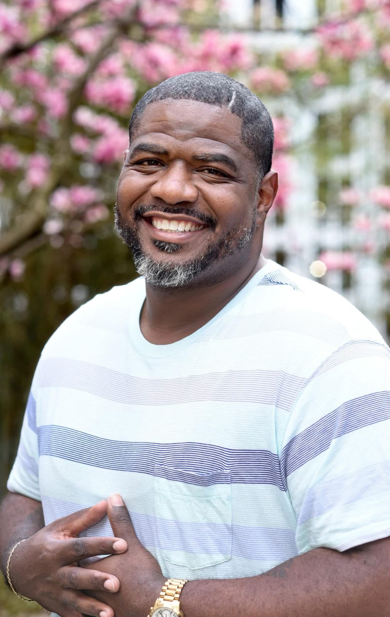 A smiling man with a beard poses outside, hands on chest. Behind him, pink flowers and a white wooden fence.