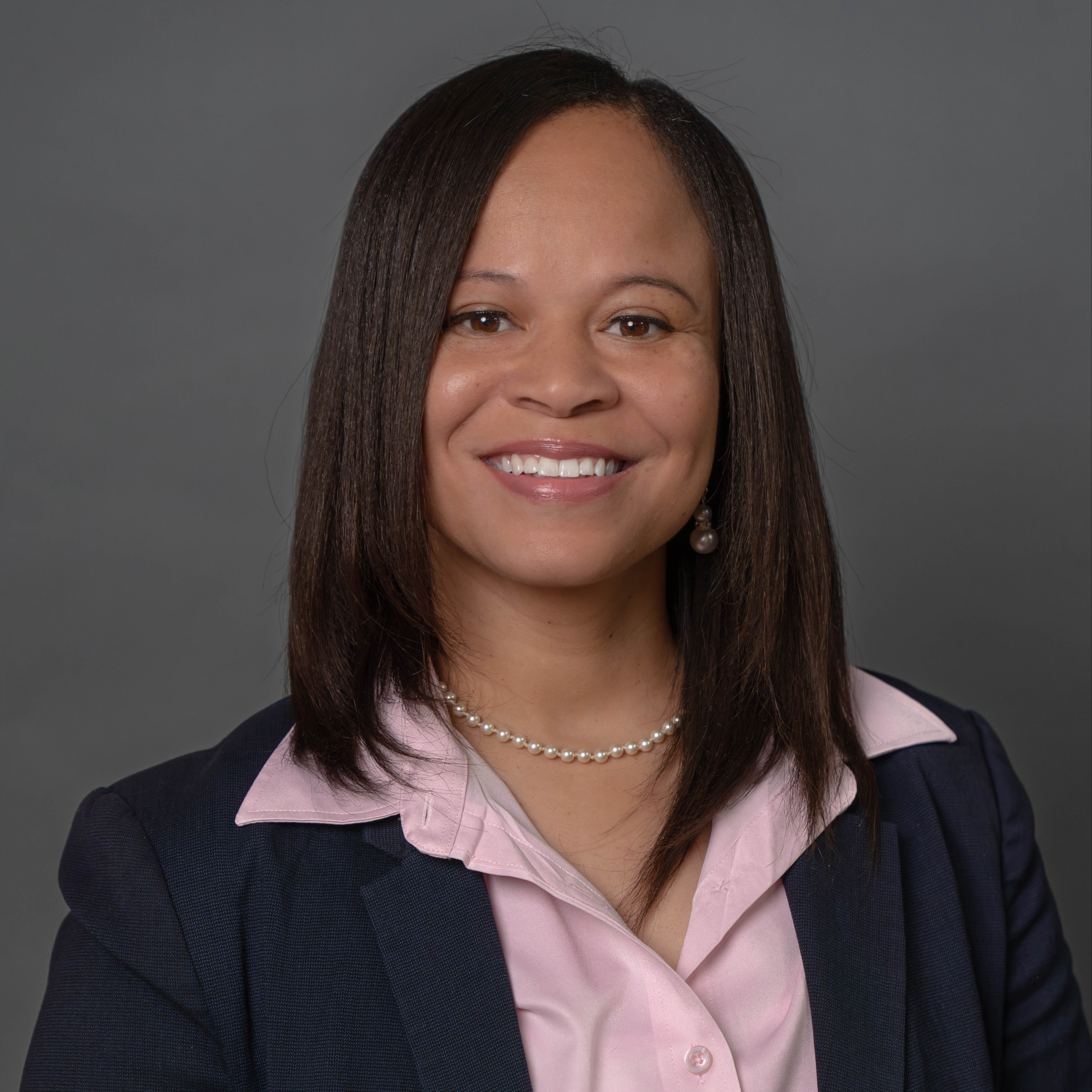 Woman in pink shirt with navy blazer and pearl necklace stands against gray background. She smiles and looks at camera.
