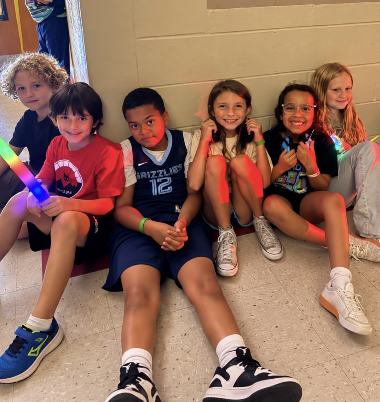 Six children sit on the floor, some with face paint, holding glow sticks. A beige wall is behind them.