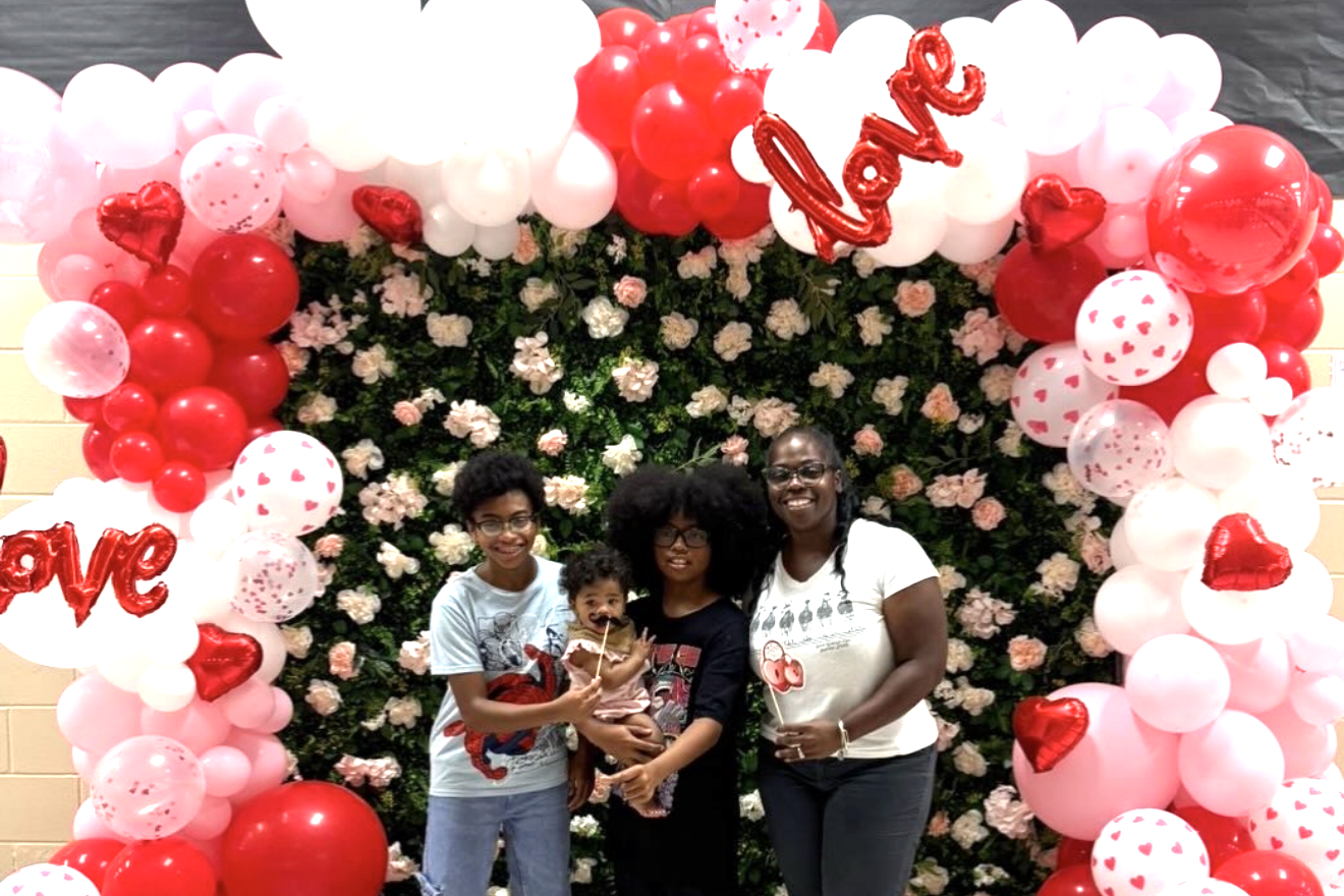 Three individuals standing before a pink and white balloon arch decorated with red hearts and white polka dots.