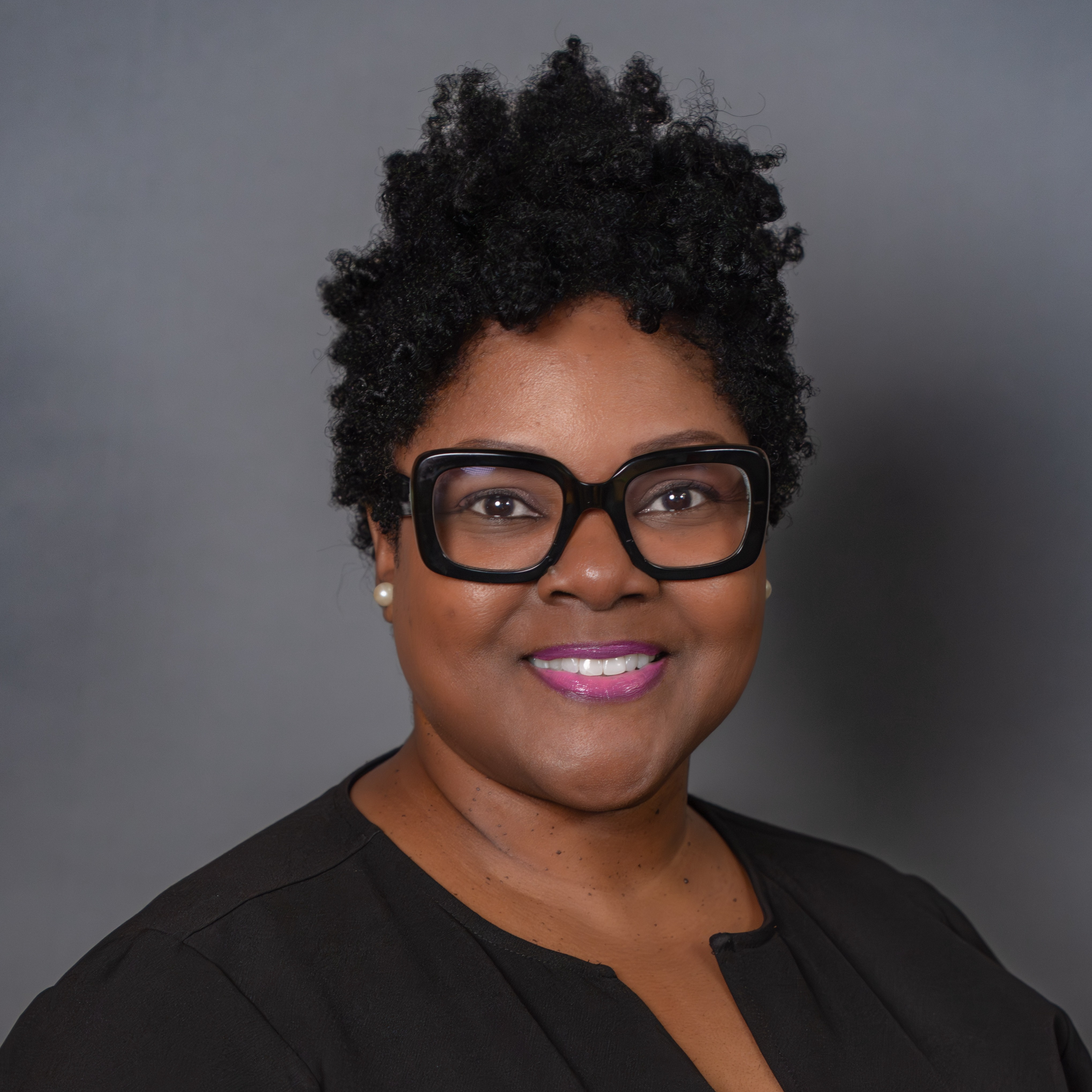 A woman in glasses and a black top smiles against a gray background. She has curly, dark hair.