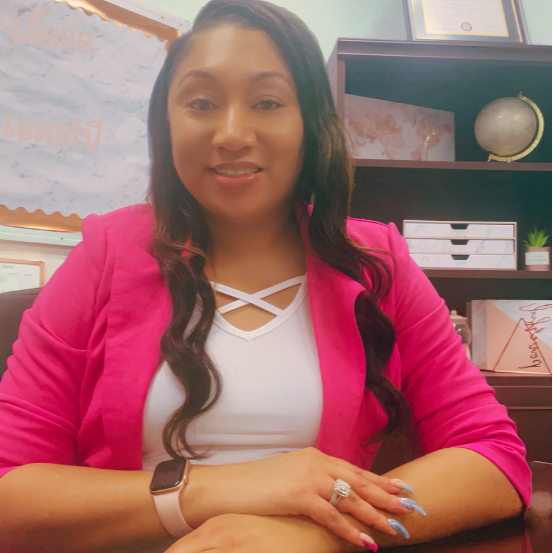 Woman in pink blazer and white top sits at a desk with hands on the table, smiling.