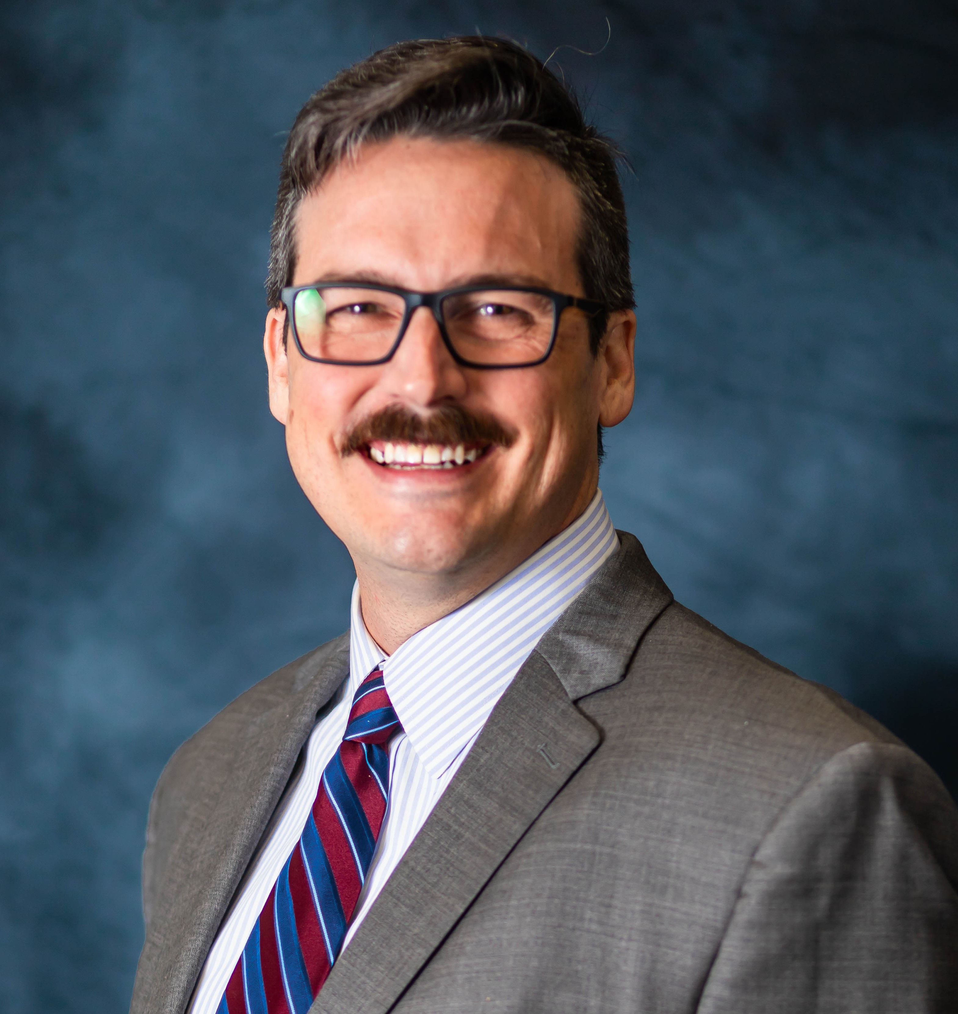 Man with glasses, mustache, and gray suit. Smiling with blue background.
