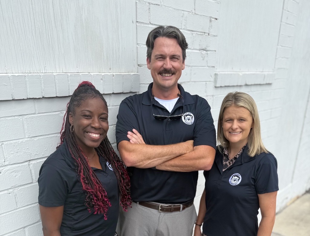 Three people in matching black shirts standing in front of a white building with a brick wall.