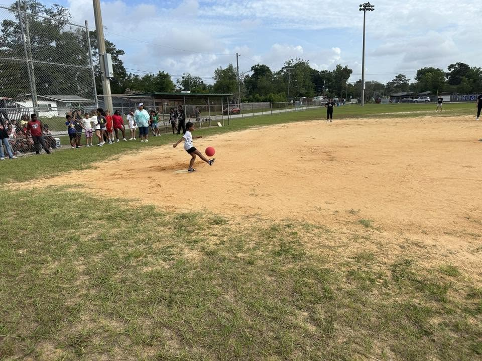 A child playing kickball on a dirt field with grass, several people watching from the sidelines.