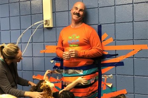 A jovial man, adorned in orange and blue, stands between two women while a group of children help him from being stuck to a wall with tape.