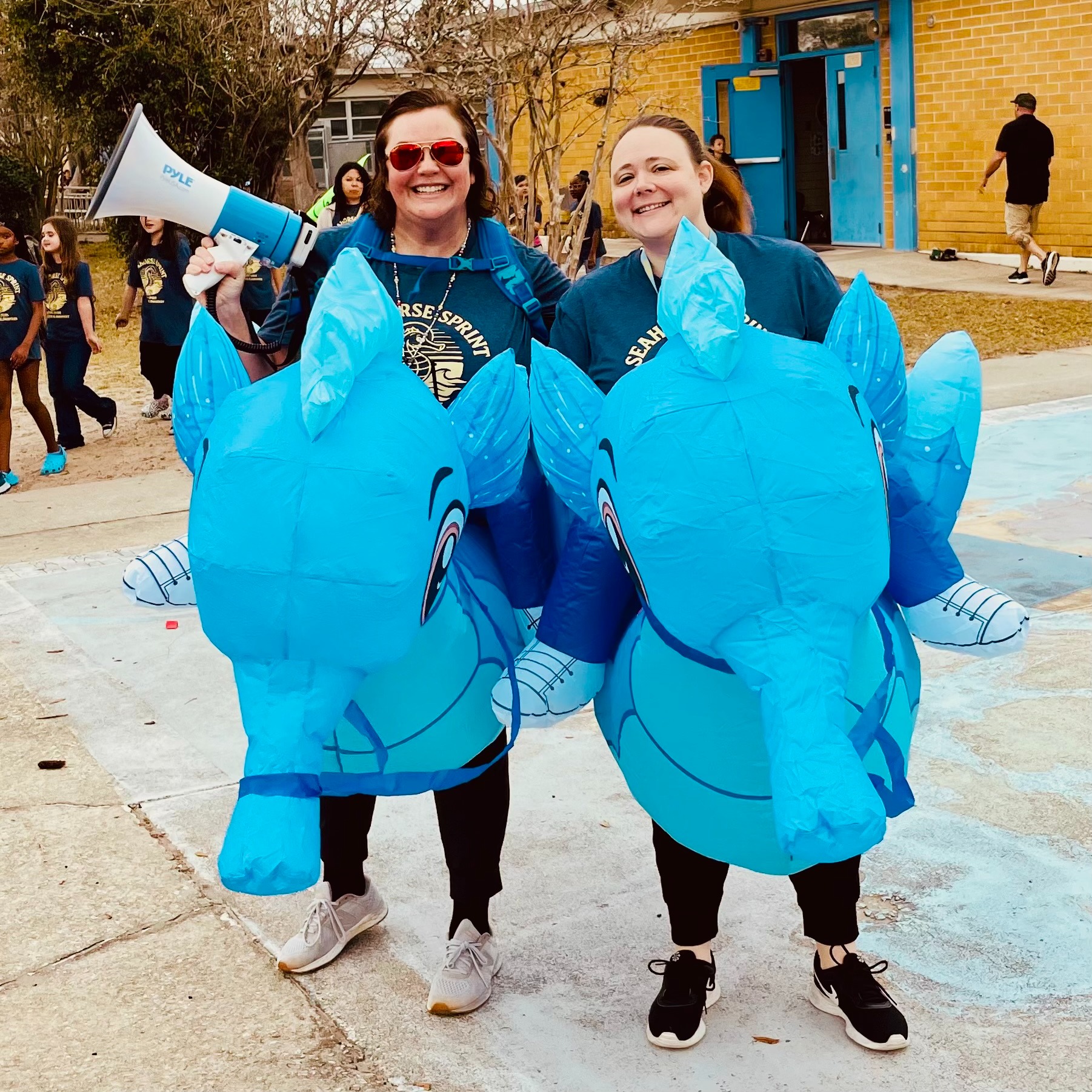 Two adults dressed as blue elephants, one holding a megaphone, standing outside a yellow building.