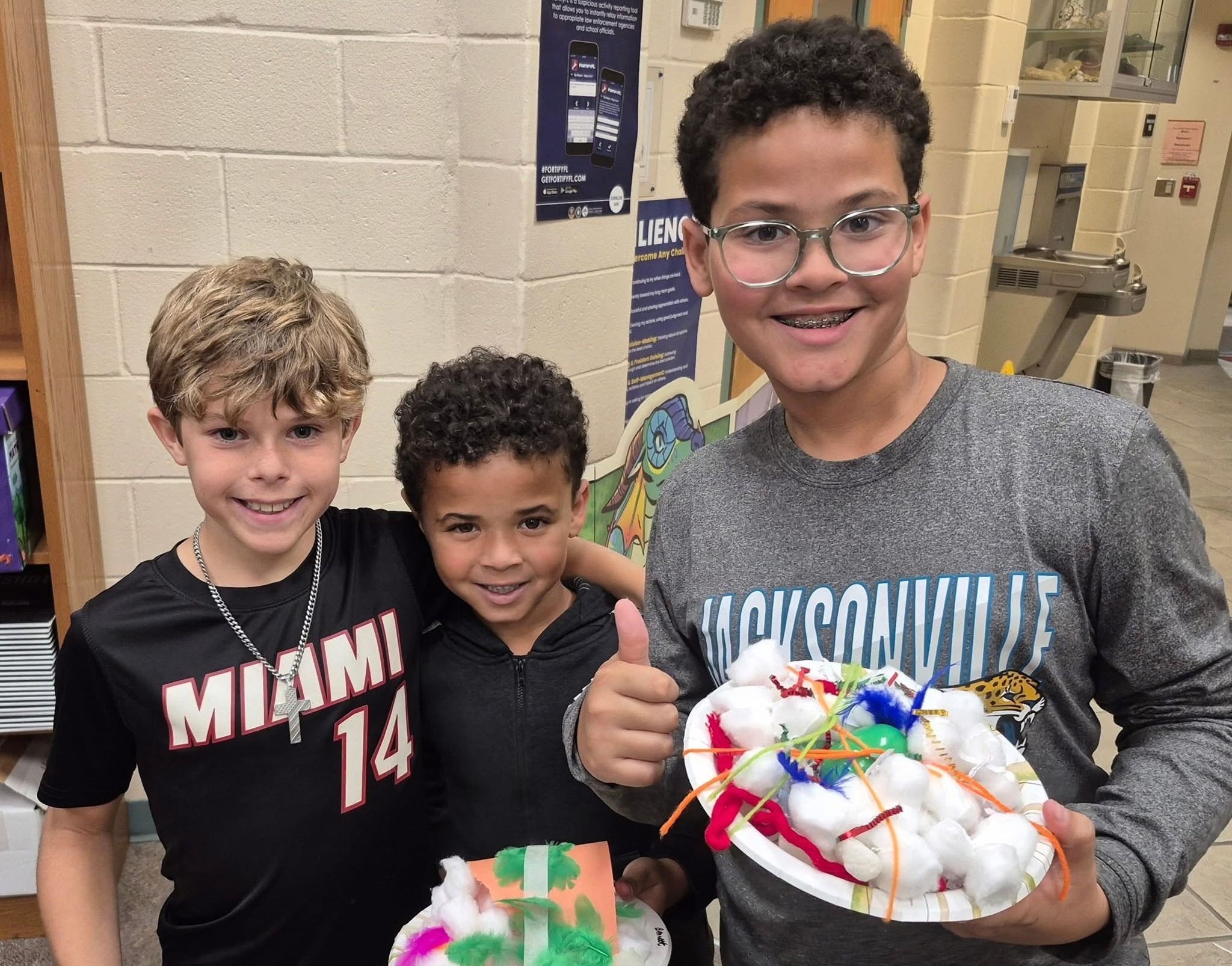 Three young boys holding art projects in plates, standing together and smiling. One boy gives a thumbs up.