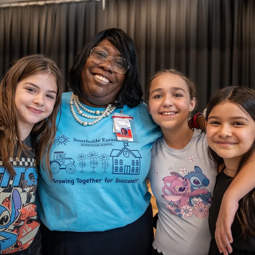 Three girls and an adult woman stand close together, smiling. The woman has a necklace and glasses.